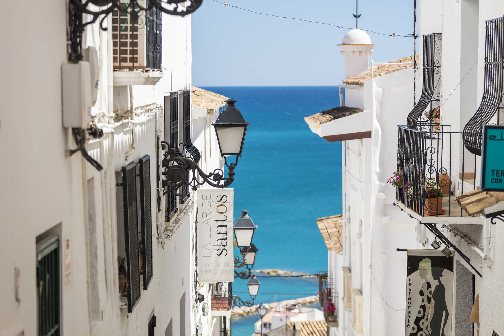 View of a narrow street in Altea's old town, featuring white-washed facades, traditional wrought-iron lanterns, and a glimpse of the turquoise Mediterranean Sea.