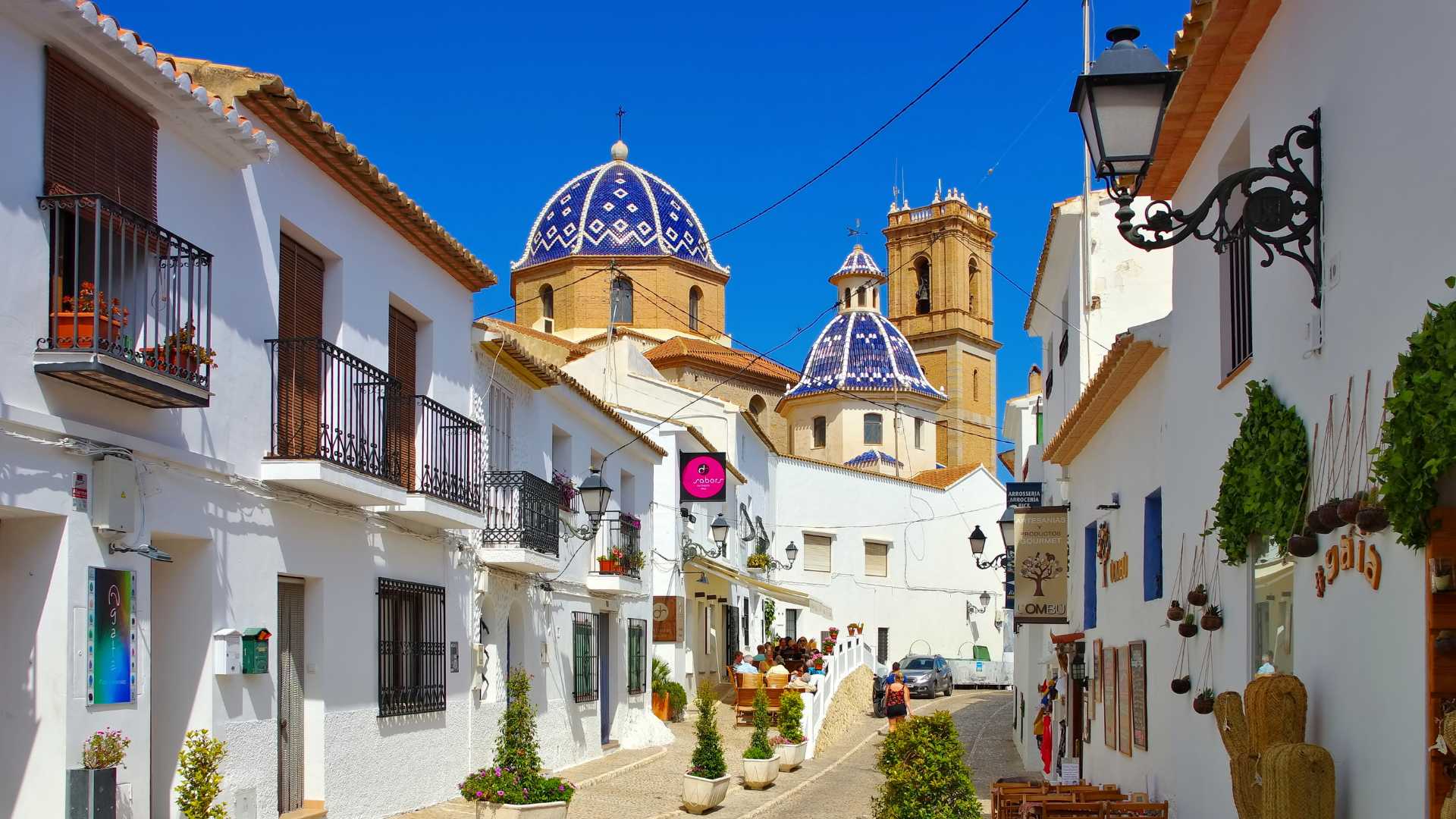 View of Altea's old town featuring narrow cobbled streets, traditional white-washed houses, and the iconic blue-and-white tiled church domes against a clear sky.