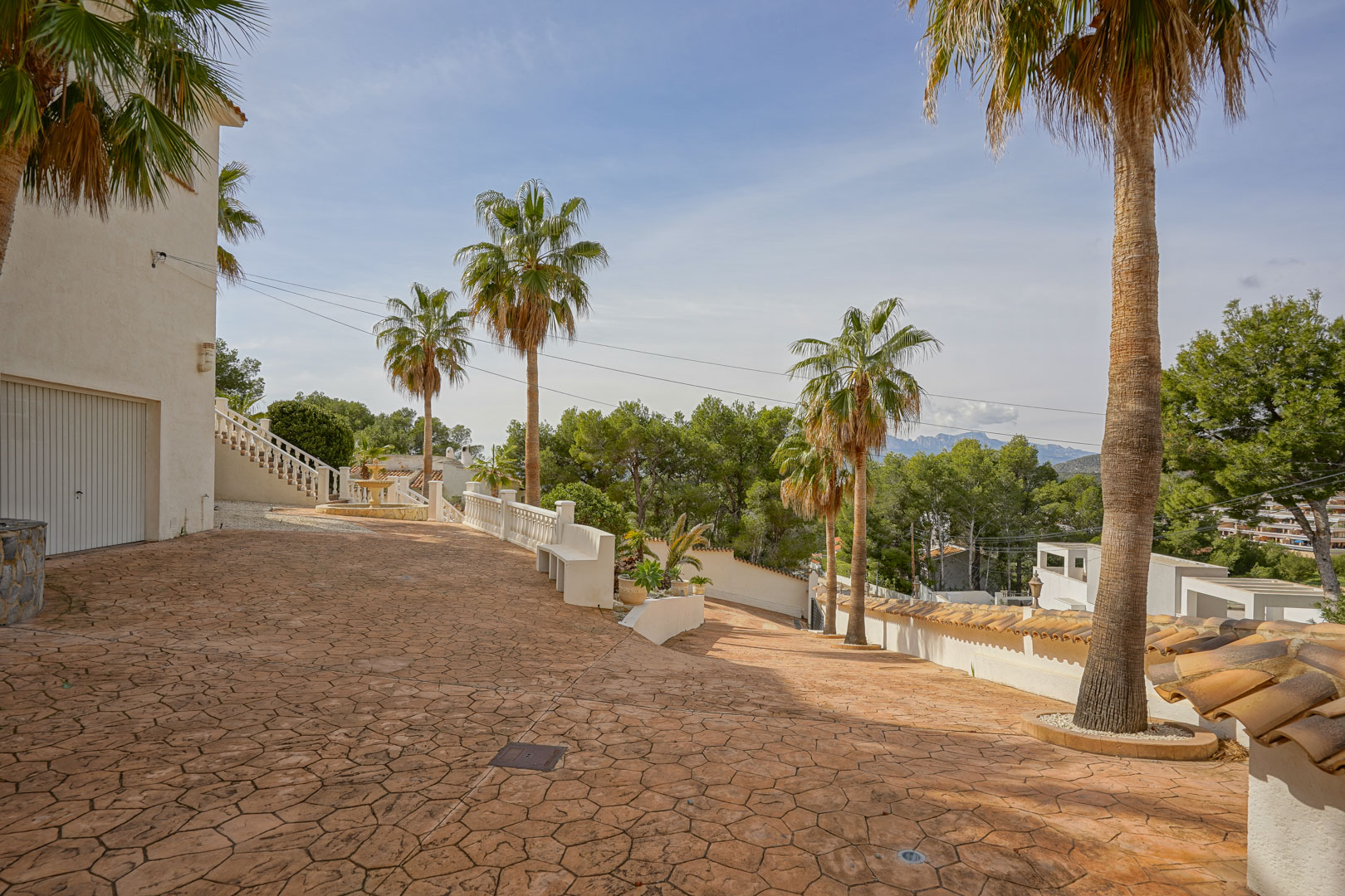 Villa entrance featuring a spacious stamped concrete driveway, mature palm trees, and mountain views. The area includes garage access and classic white balustrades.