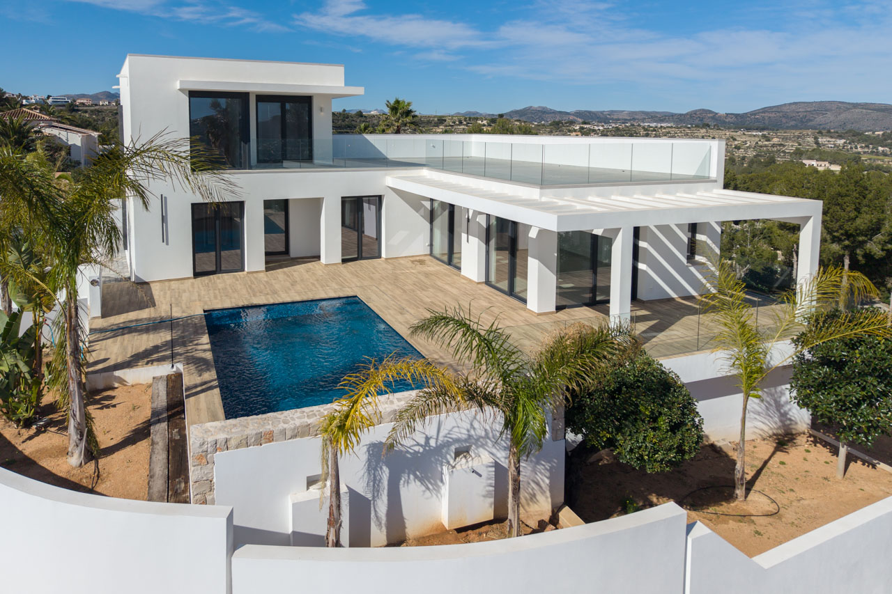 Aerial view of modern villa with blue pool, wood deck, and glass railing in Benimeit, Moraira.