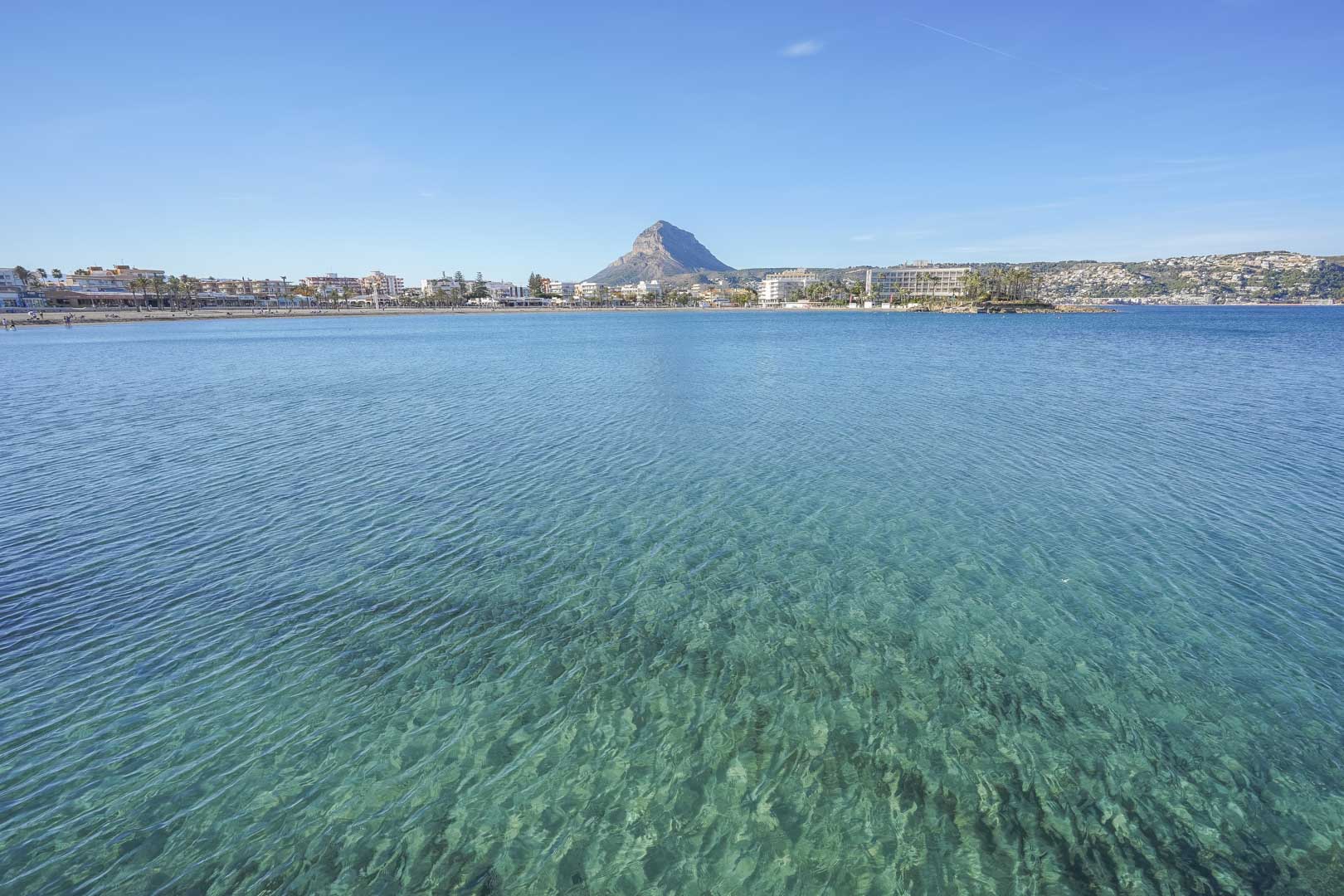 Panoramic view of the Jávea bay featuring crystal-clear turquoise waters, the urban coastline, and the prominent Montgó mountain in the background.