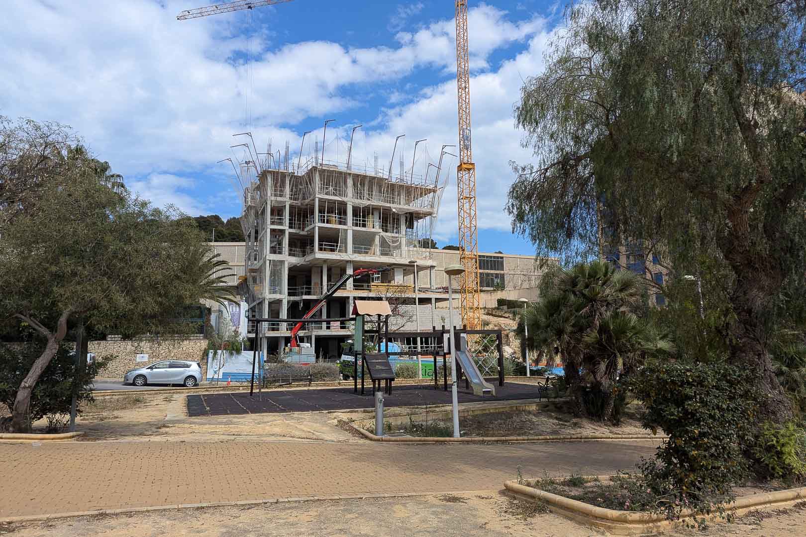 Current construction progress in Calpe, featuring the reinforced concrete structure and onsite cranes adjacent to a local park and playground area.