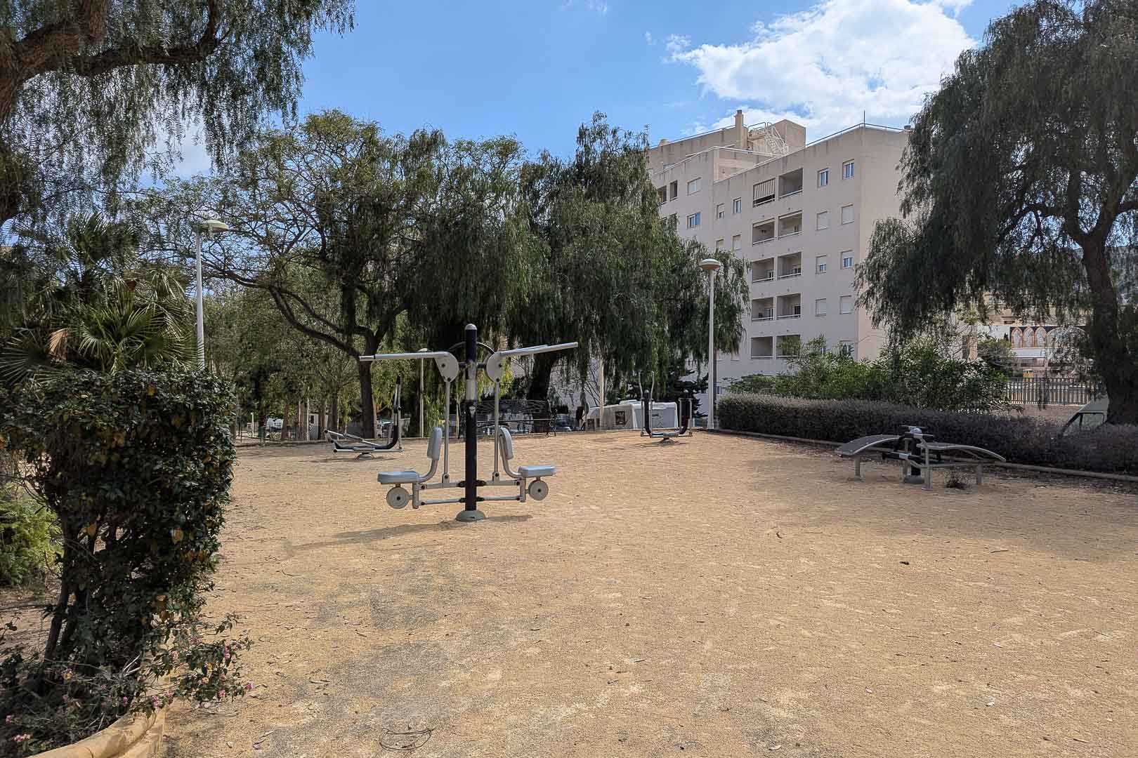 Outdoor fitness area featuring bio-healthy exercise equipment, set within a landscaped park with mature trees and nearby residential architecture.