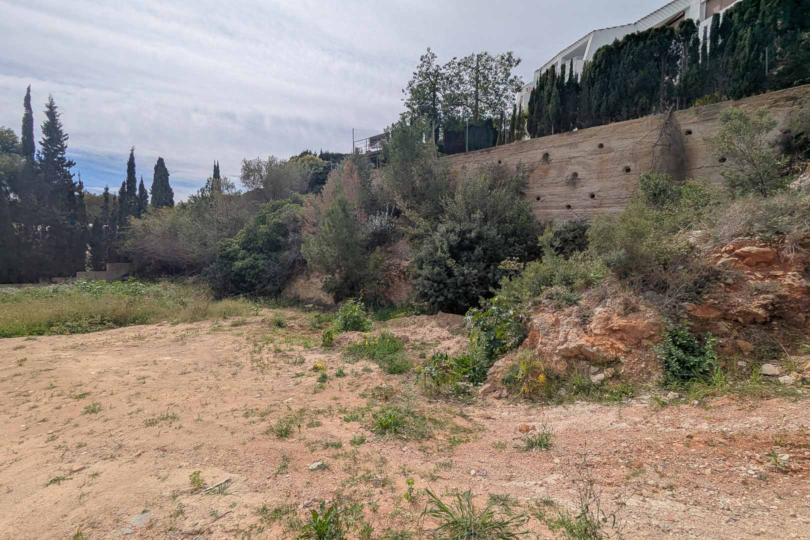 Elevated plot in La Corona featuring a concrete retaining wall and native Mediterranean vegetation. The site is ready for a custom-built villa project.