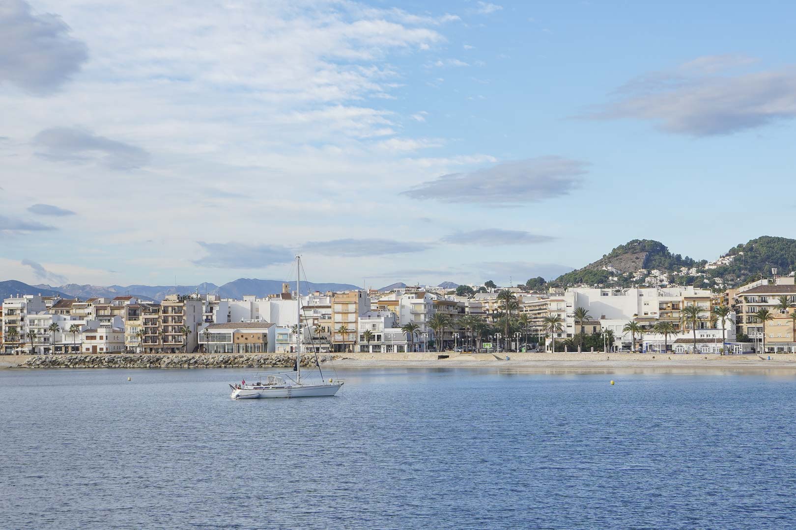 Panoramic view of the Jávea port area featuring a sailboat anchored in the bay, coastal architecture, and the surrounding Mediterranean mountains.