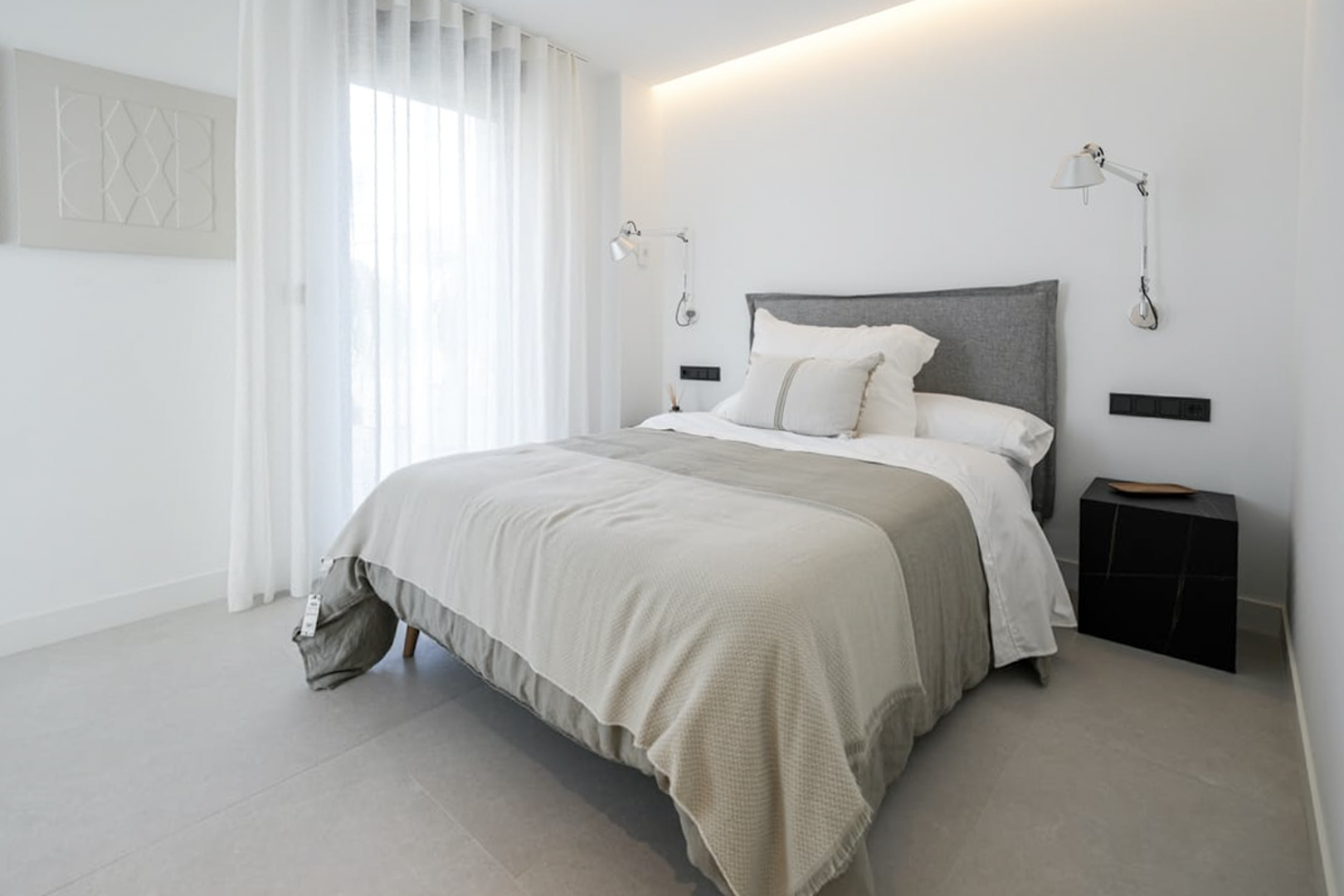 Bright bedroom featuring a double bed, gray upholstered headboard, and adjustable wall lamps. Porcelain tile flooring.