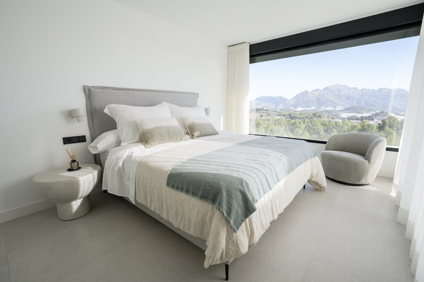 Bright bedroom with a double bed, gray armchair, and mountain views through a large window.