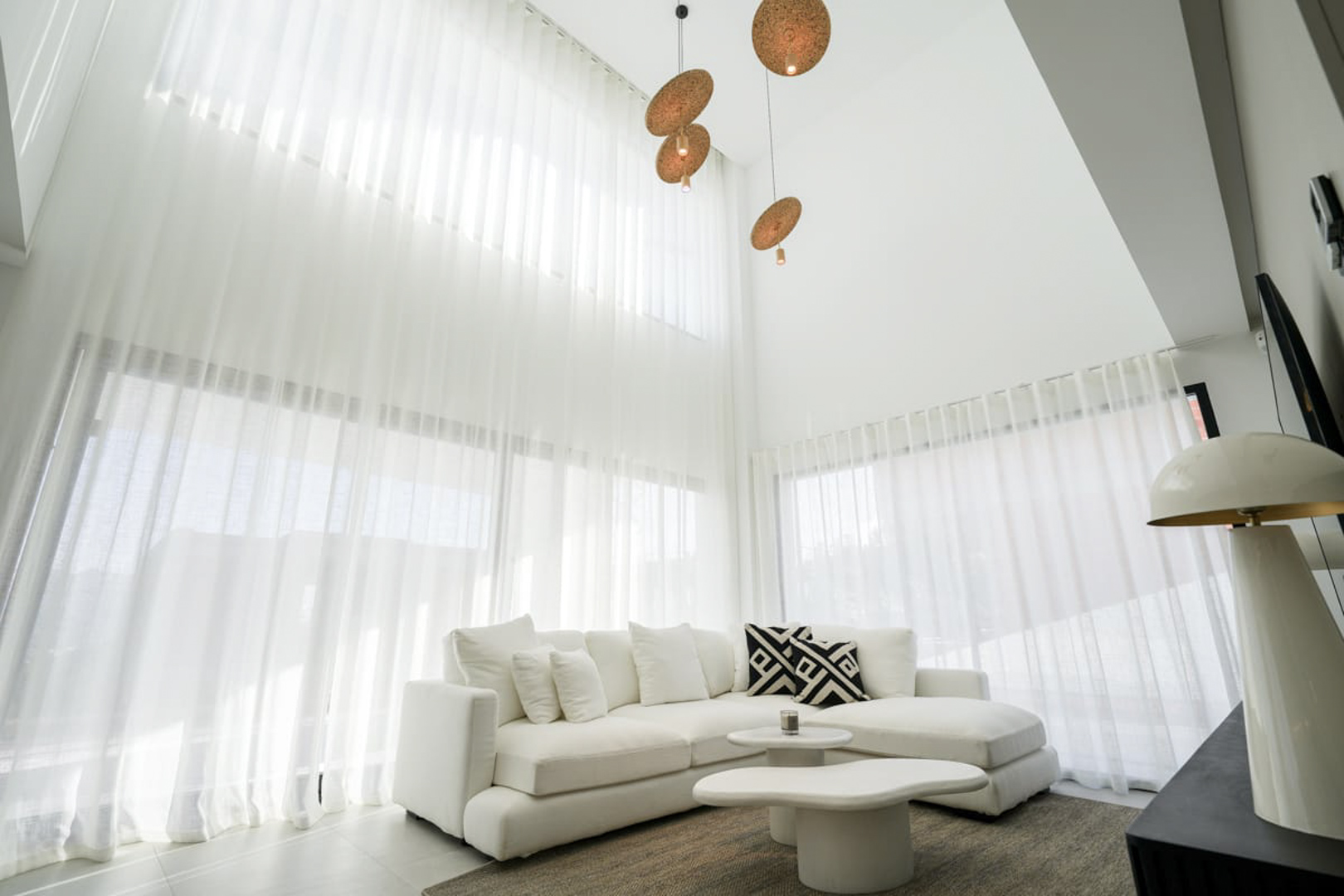 Bright living room featuring white curtains, sectional sofa, and designer pendant lights, creating a modern, minimalist ambiance.
