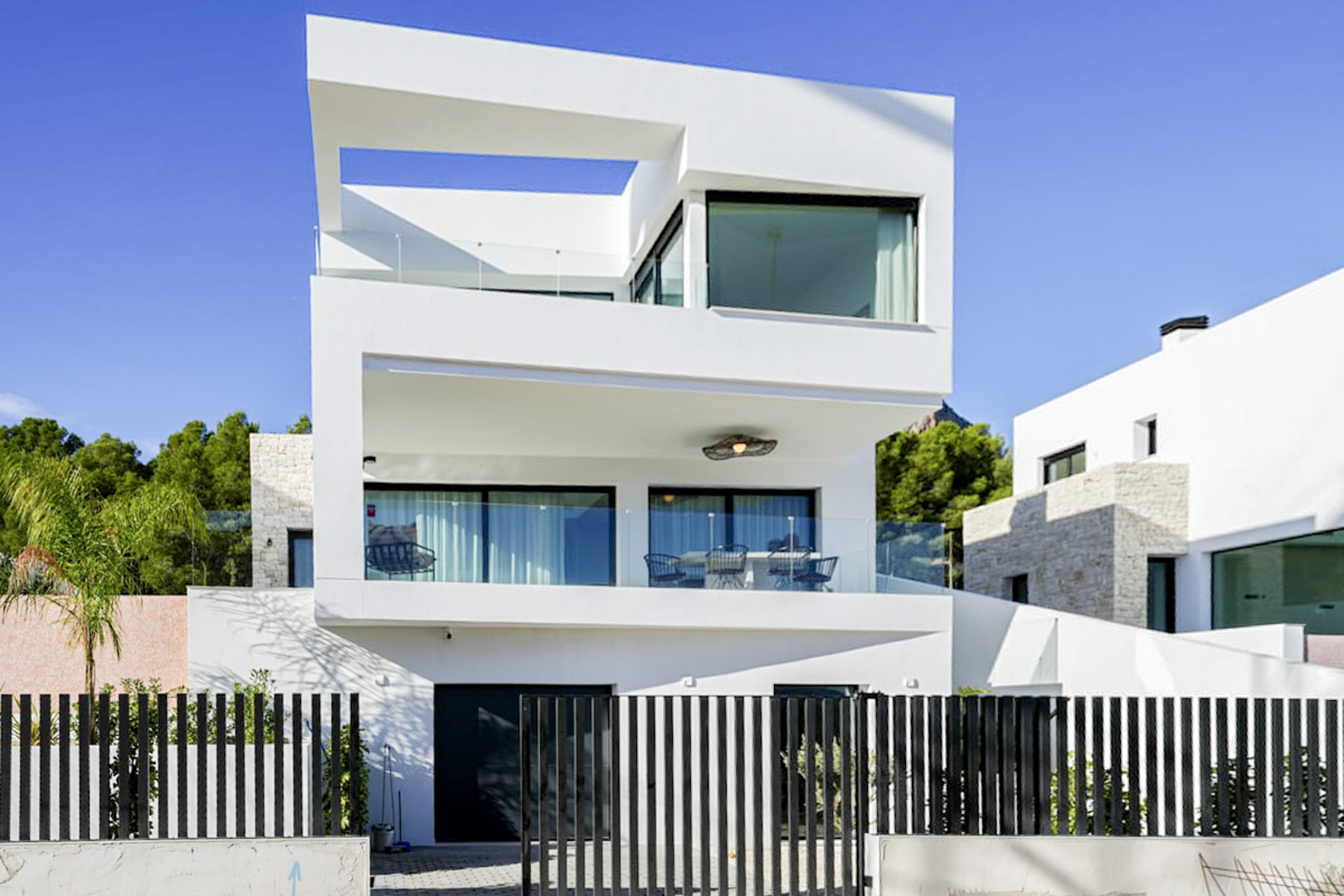 Exterior view of a modern three-story villa with balconies, glass railings, and a black slat fence.