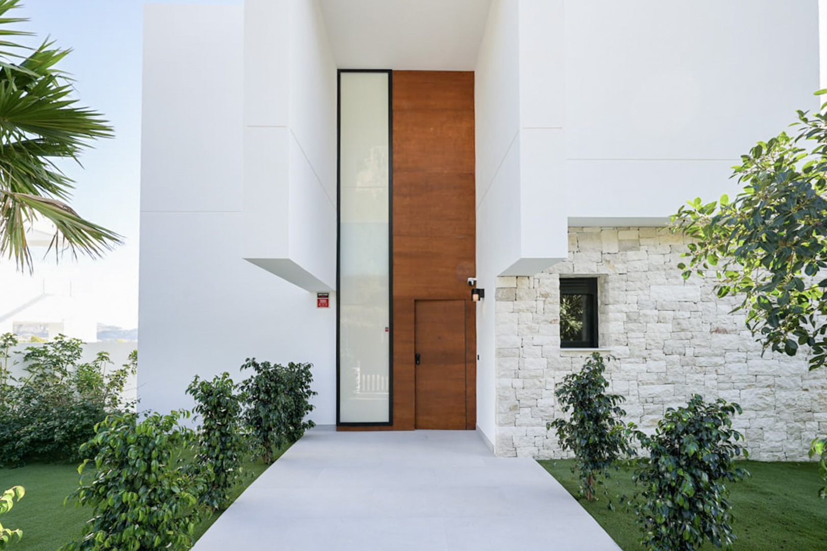 Modern facade with a wooden door and light stone accents, surrounded by greenery.