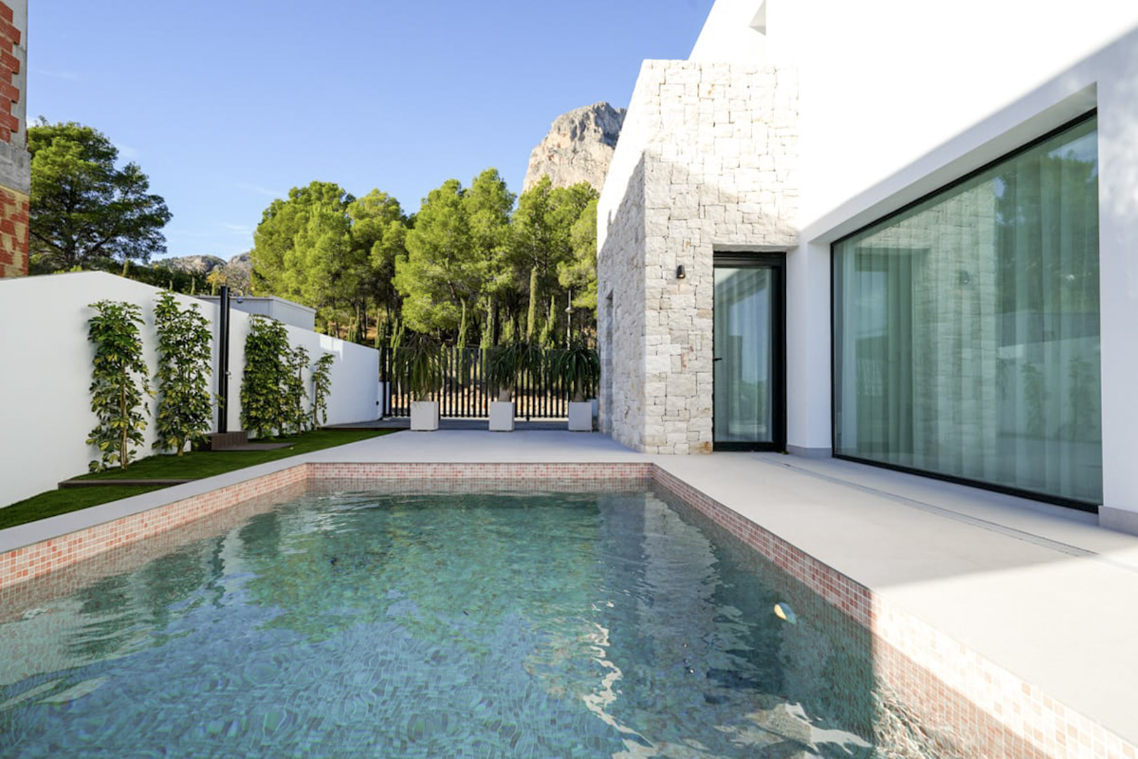 View of the private pool with mosaic trim and white stone facade, surrounded by lush greenery.