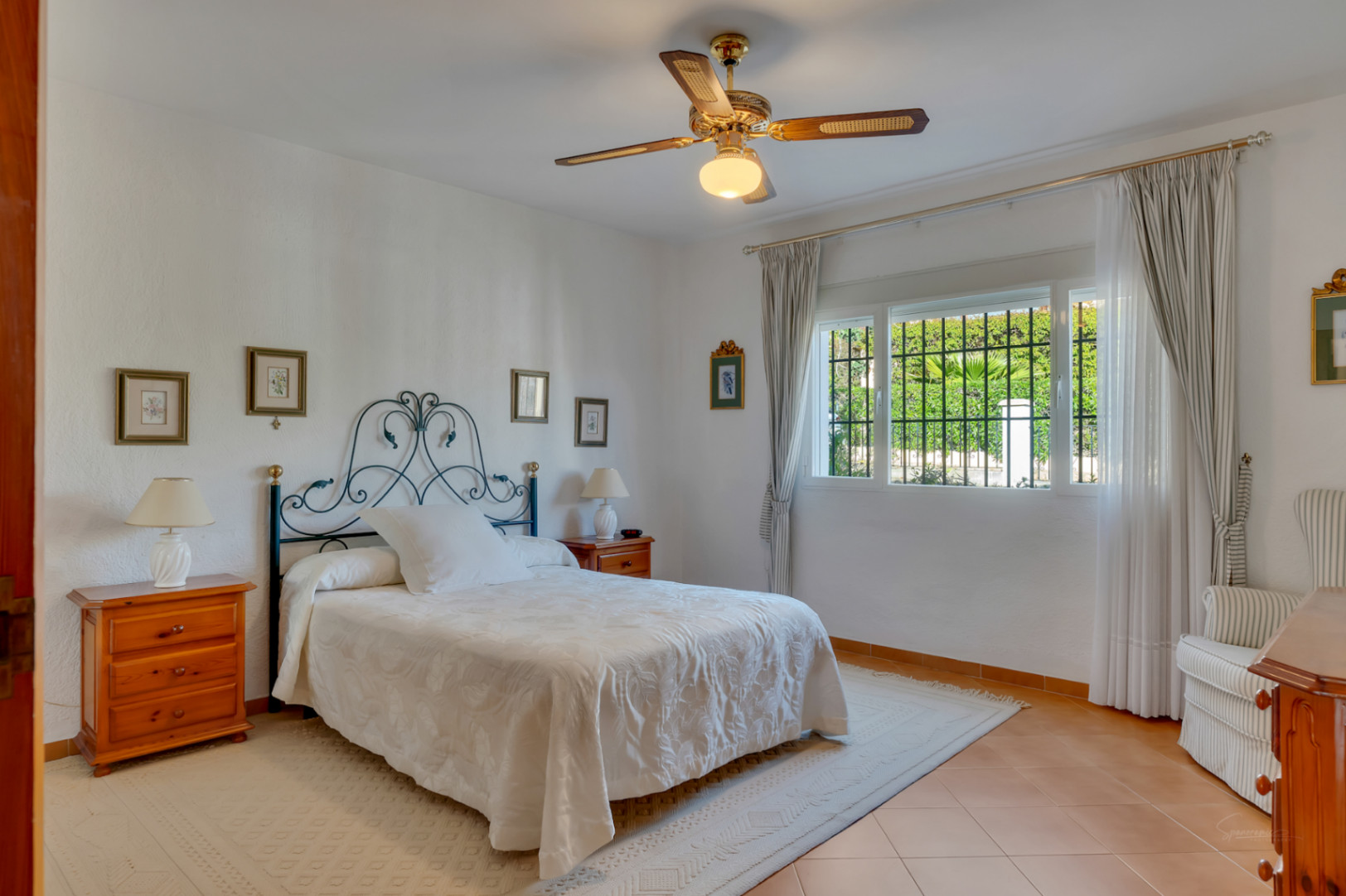 Bright bedroom featuring a wrought iron headboard, terracotta tile flooring, and a ceiling fan. Large windows with security bars offer natural light and views of the garden.