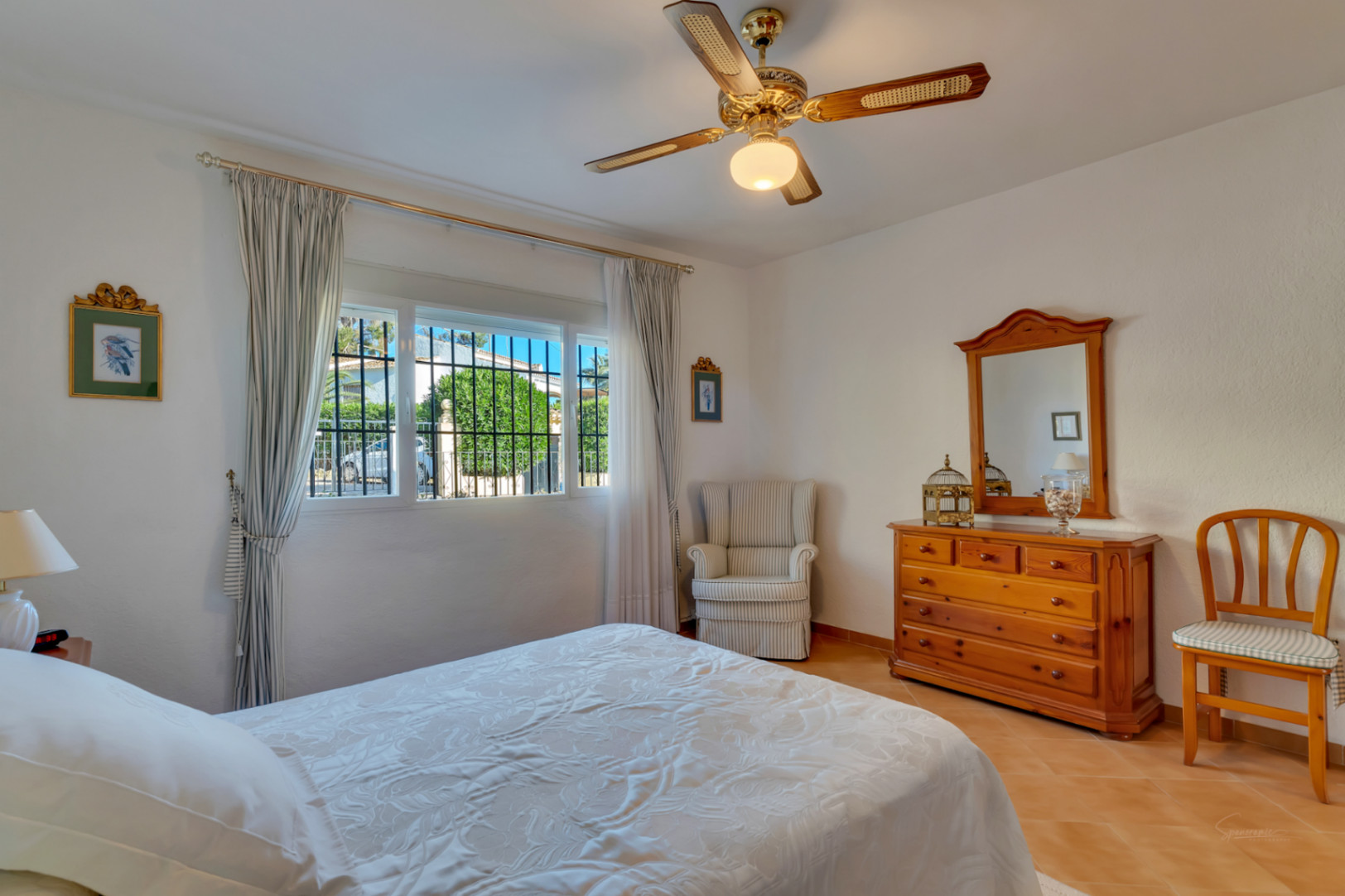 Bright bedroom featuring terracotta tile flooring, traditional wooden furniture, a ceiling fan, and a large window overlooking the garden.