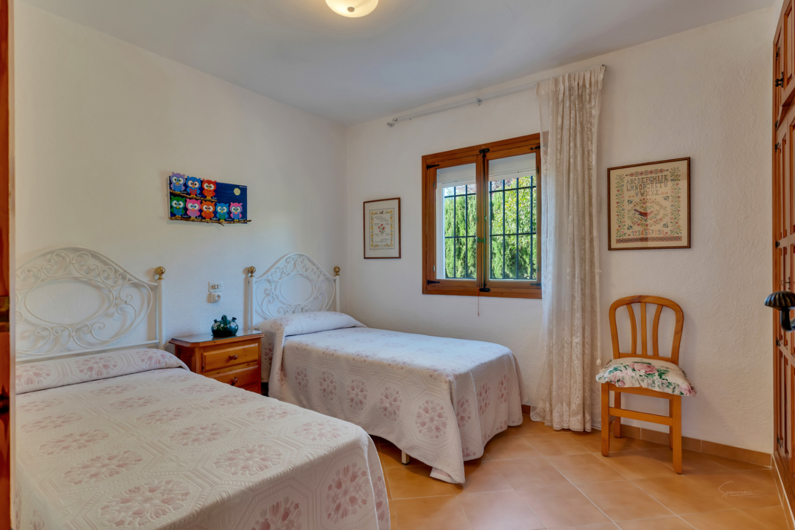 Bright bedroom featuring twin beds with white wrought-iron headboards, terracotta tile flooring, and a traditional wooden window frame.