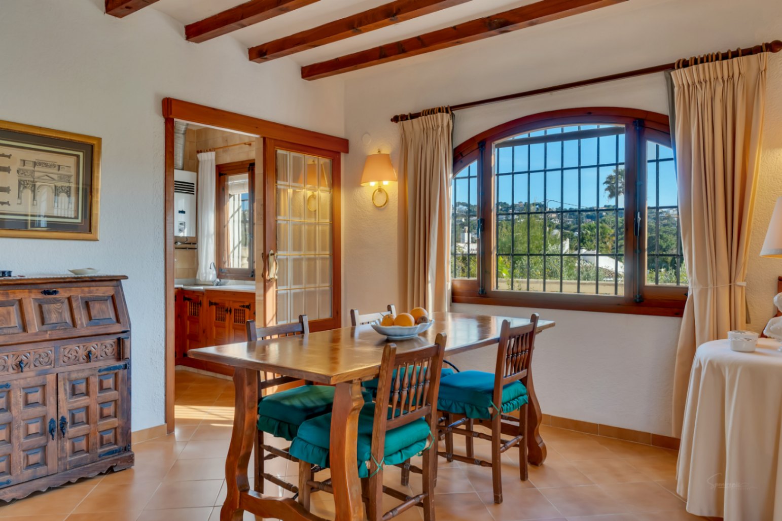 Bright dining area featuring exposed wooden ceiling beams, terracotta flooring, and an arched window. A sliding glass door connects the space to the traditional kitchen.