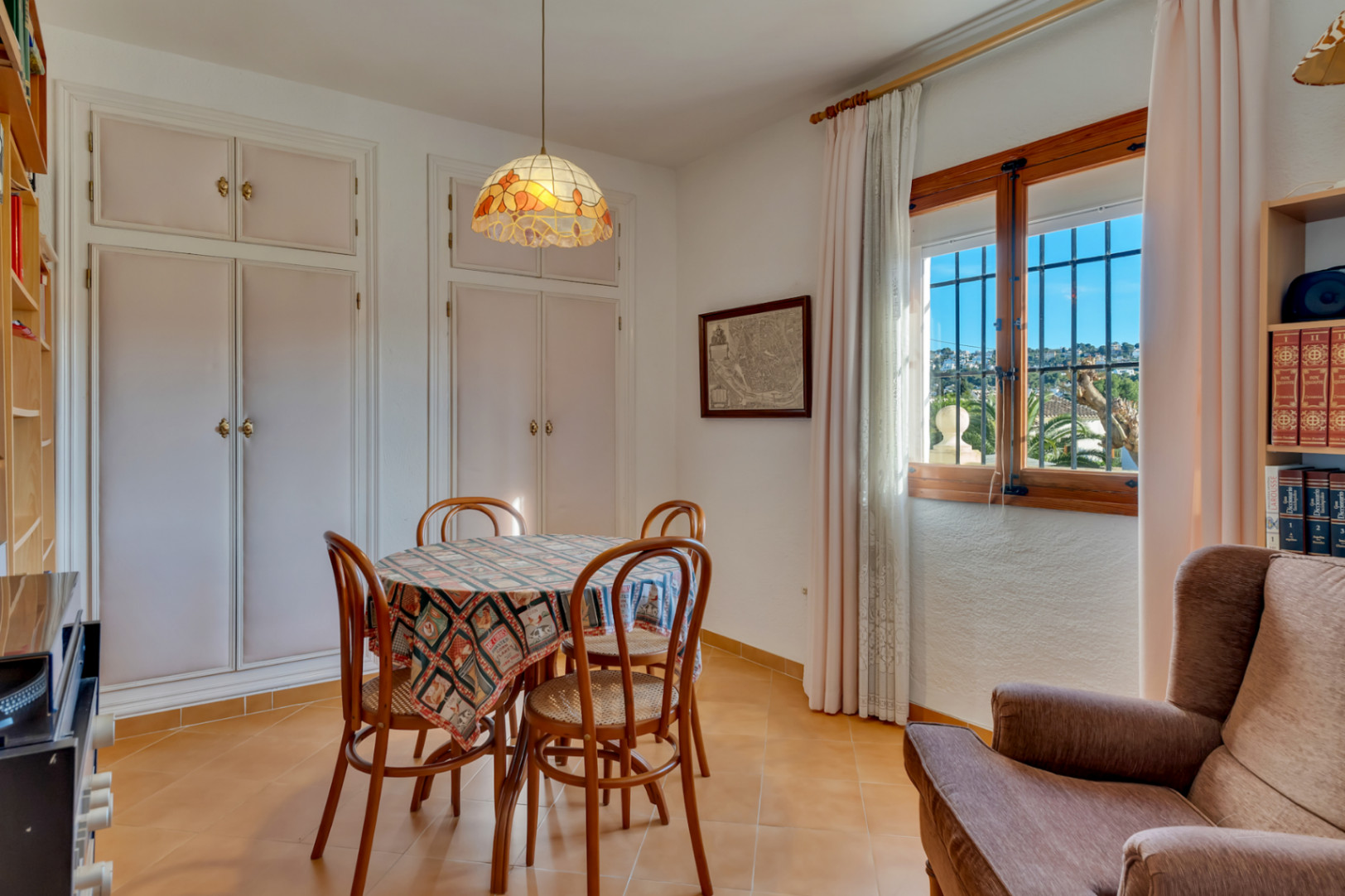 Bright dining space featuring white built-in wardrobes, a round table with wooden chairs, and terracotta tiles. Large window provides natural light and neighborhood views.