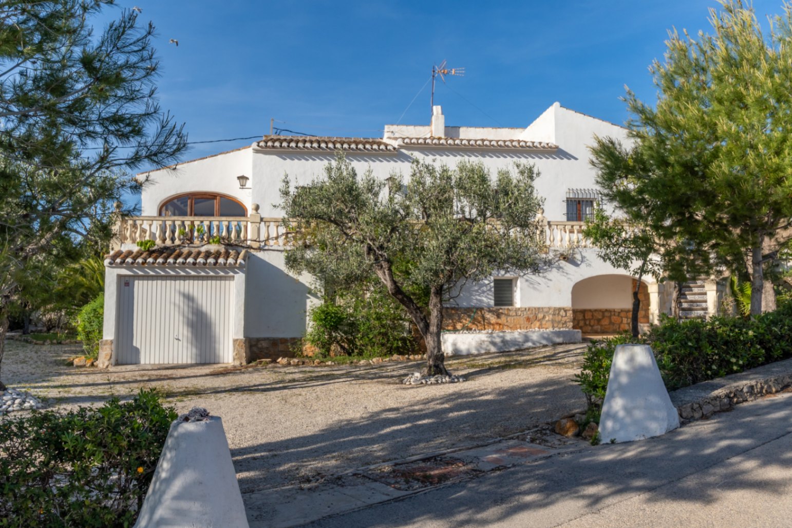 Exterior of the villa showcasing traditional white architecture, an integrated garage, a balustrade terrace, and a gravel driveway with mature olive trees.