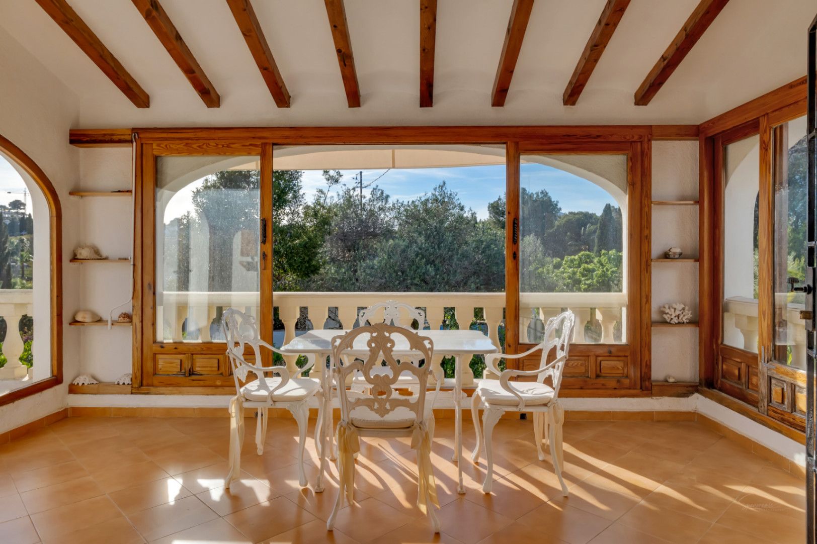 Glazed terrace featuring exposed wooden ceiling beams, terracotta flooring, and white ornate metal furniture with views of the surrounding greenery.