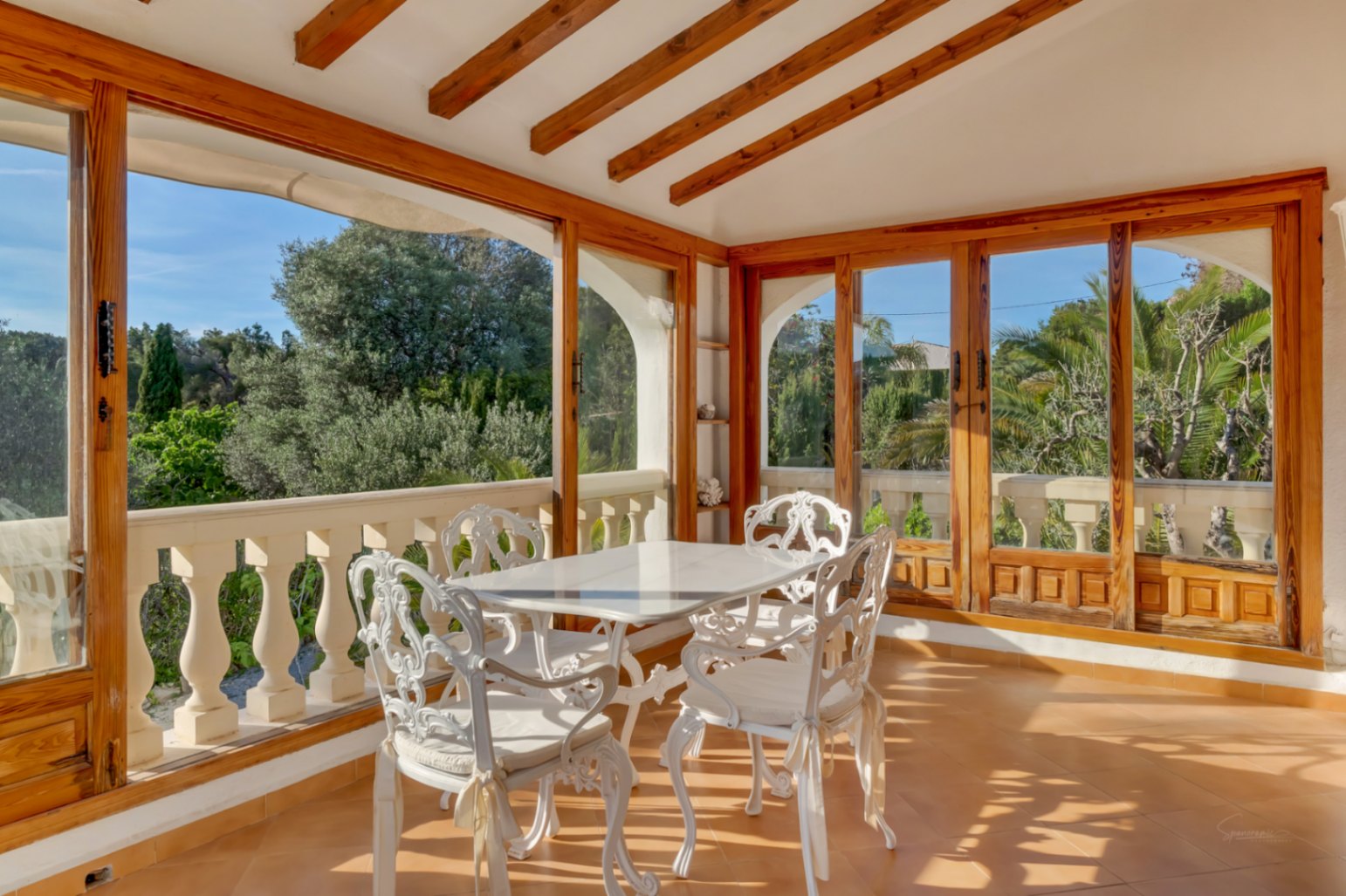 Glazed terrace featuring exposed wooden ceiling beams, white ornate metal dining furniture, and large windows overlooking the lush Mediterranean garden.