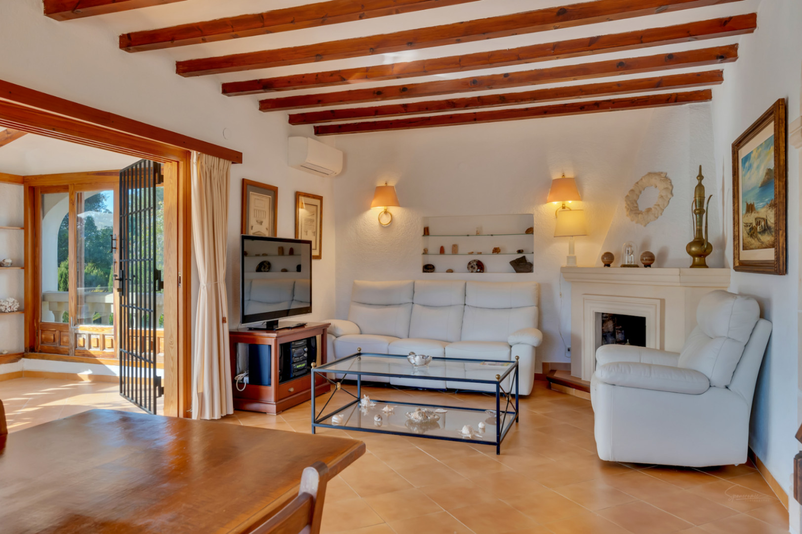 Living room featuring exposed dark wood ceiling beams, a classic fireplace, and terracotta flooring. Large glass doors provide direct access to the bright terrace.