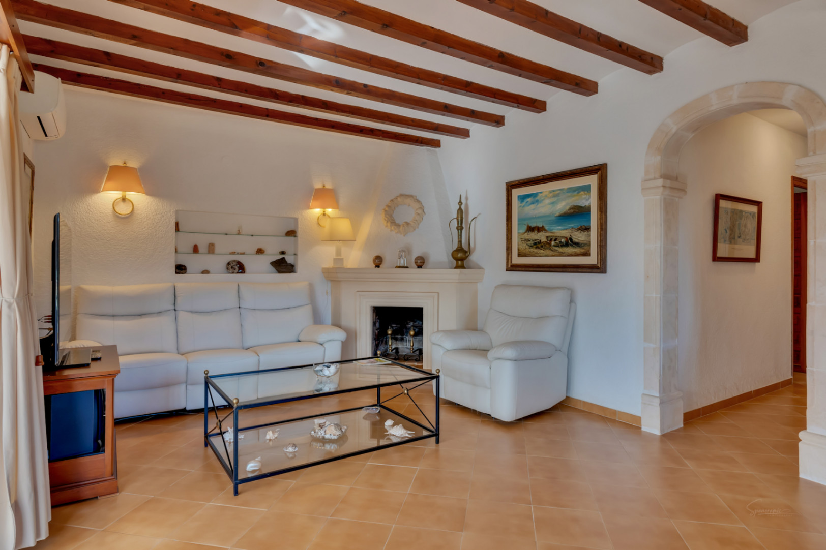 Living room featuring exposed wooden ceiling beams, a classic fireplace, and a traditional tosca stone archway. White leather seating complements the terracotta-style flooring.