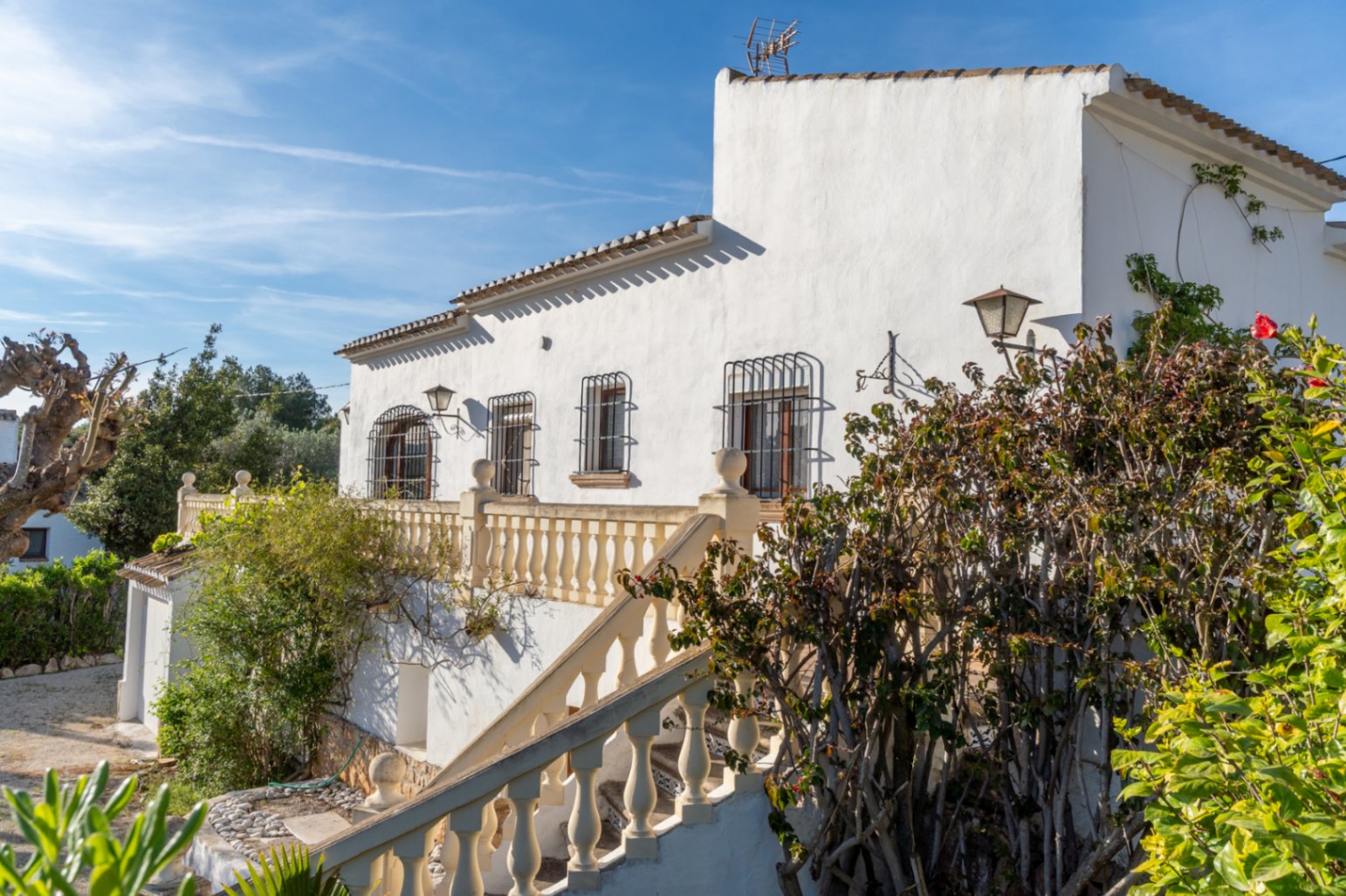 Mediterranean-style white facade featuring an exterior staircase with balustrades, windows with decorative iron bars, and mature garden greenery.