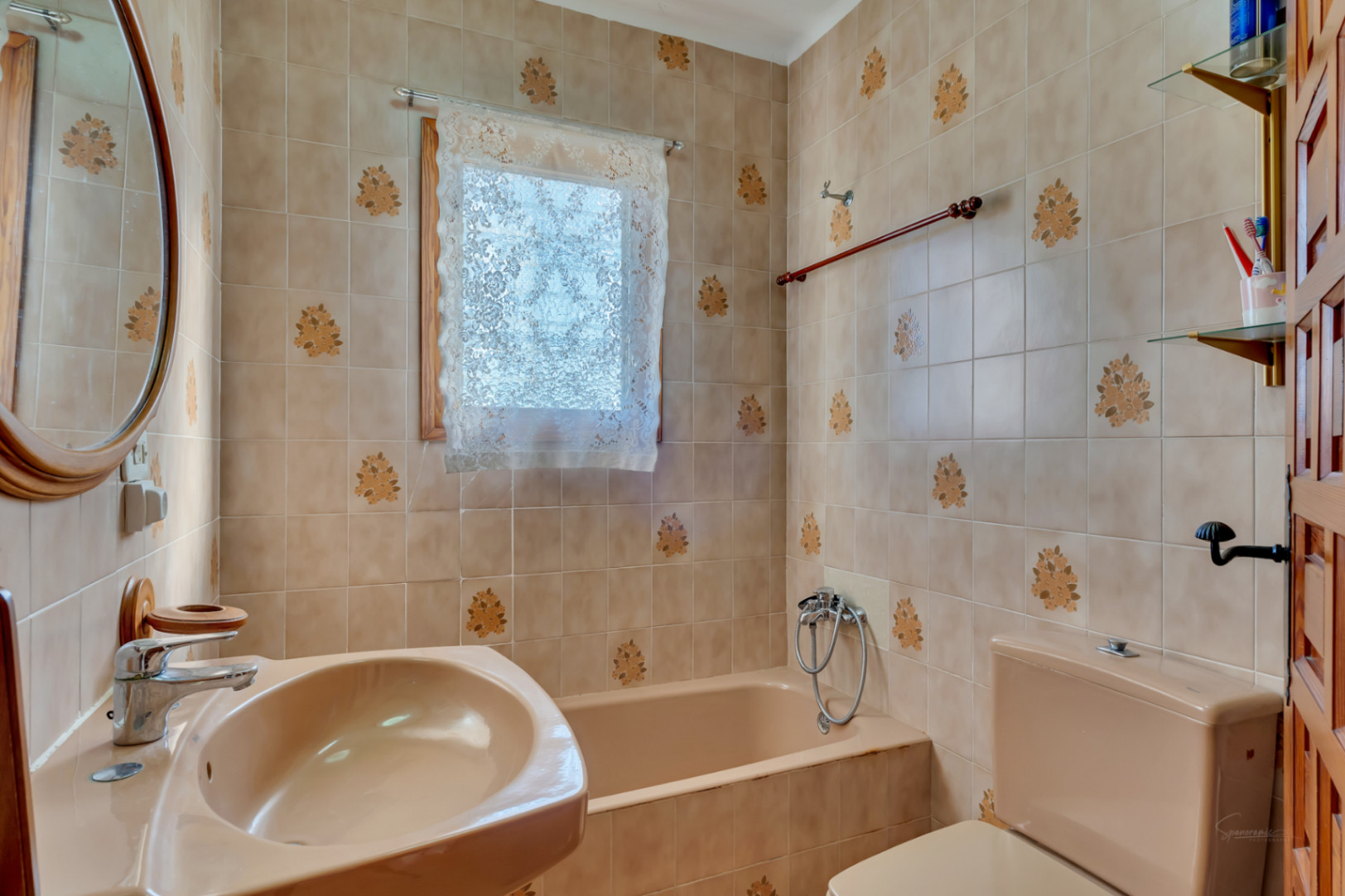 Traditional bathroom featuring beige floral tiles, a matching porcelain suite with bathtub and sink, and a wooden-framed oval mirror complemented by natural light.