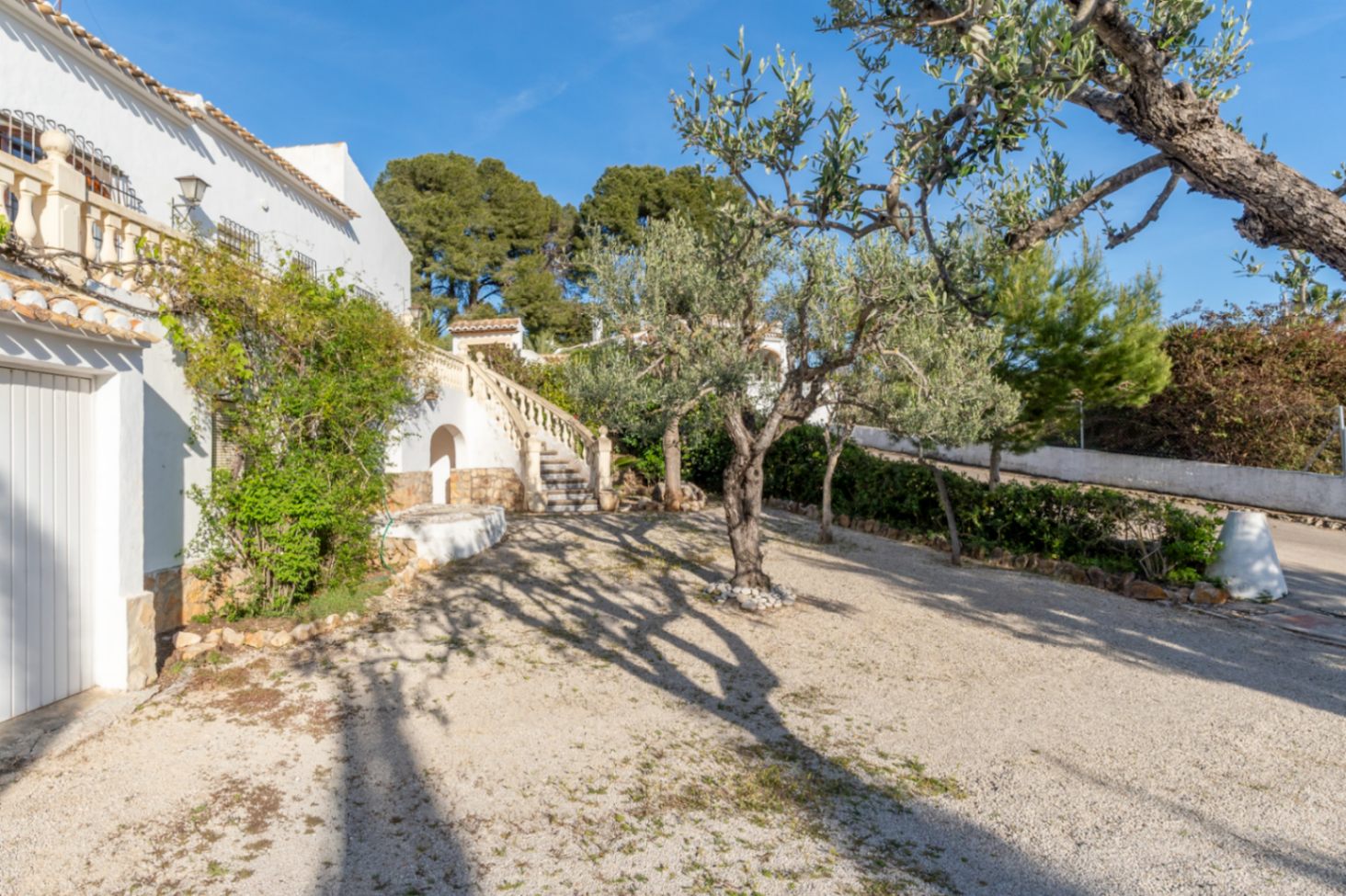 Villa exterior featuring a gravel driveway, mature olive trees, and a traditional stone staircase with balustrades leading to the upper terrace.