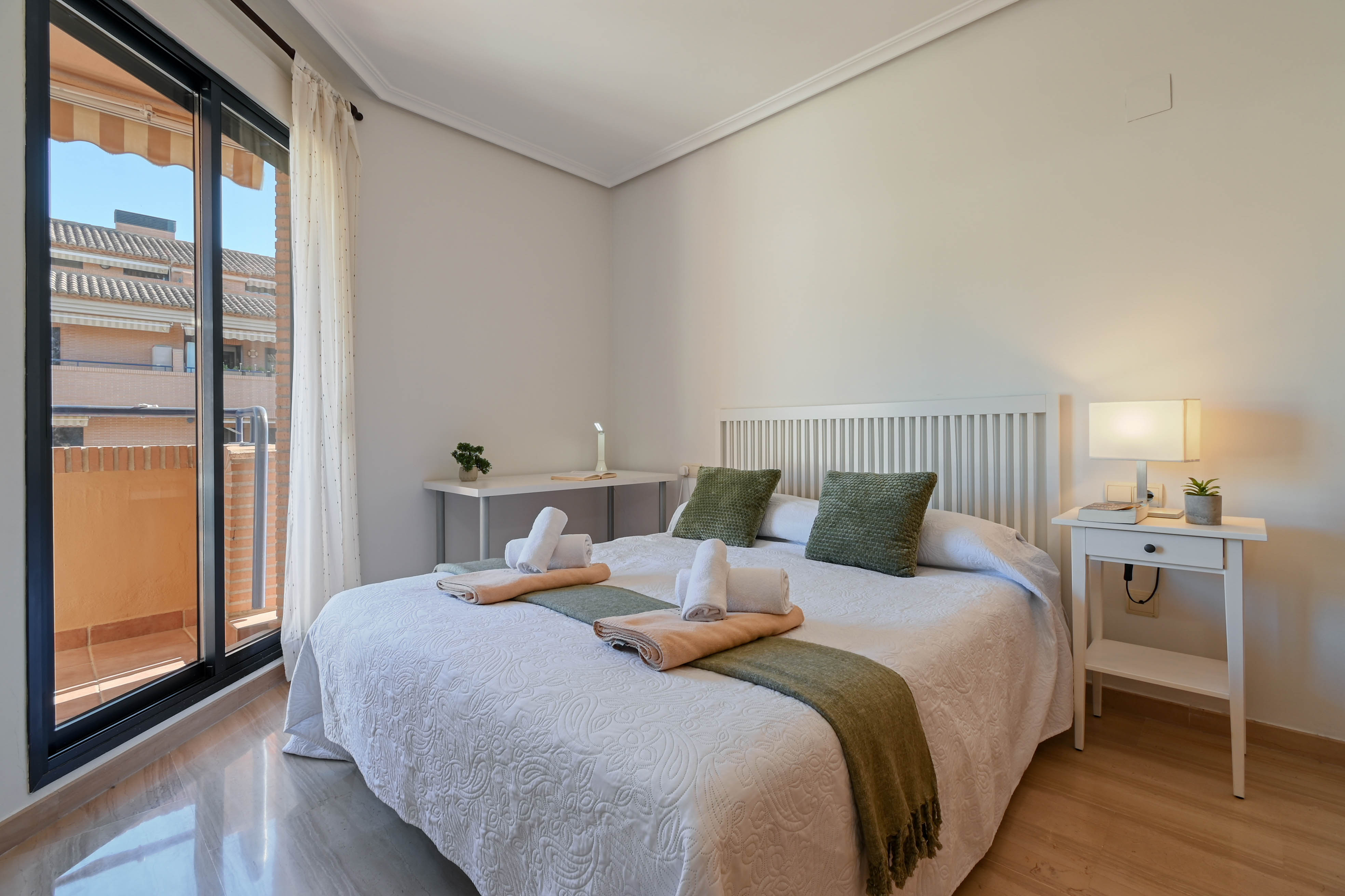 Bright bedroom featuring a double bed with a white slatted headboard, a workspace desk, and direct access to a private balcony via sliding glass doors.