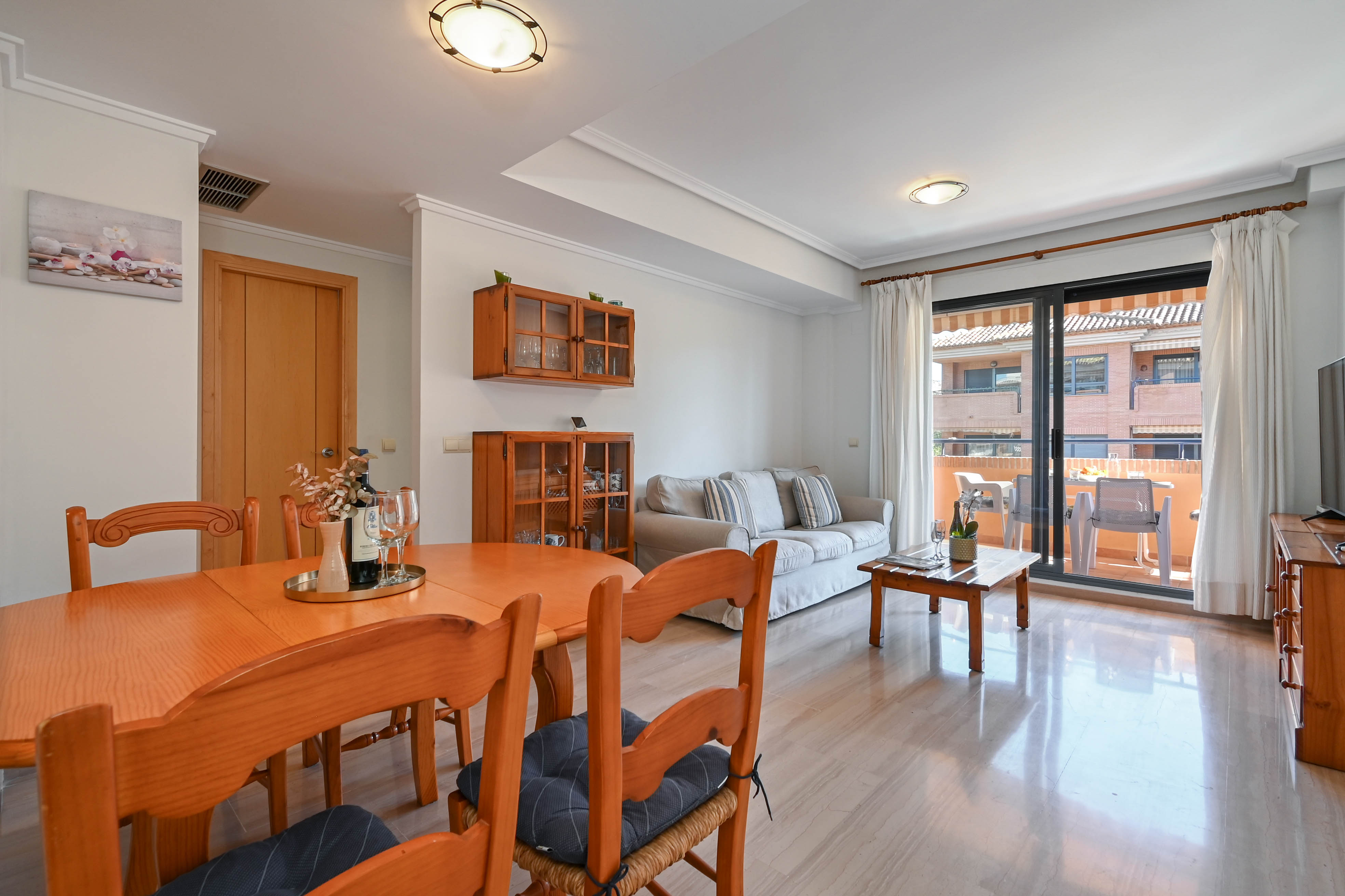 Bright living and dining area featuring wooden furniture, polished stone flooring, and large sliding doors opening onto a private furnished terrace.