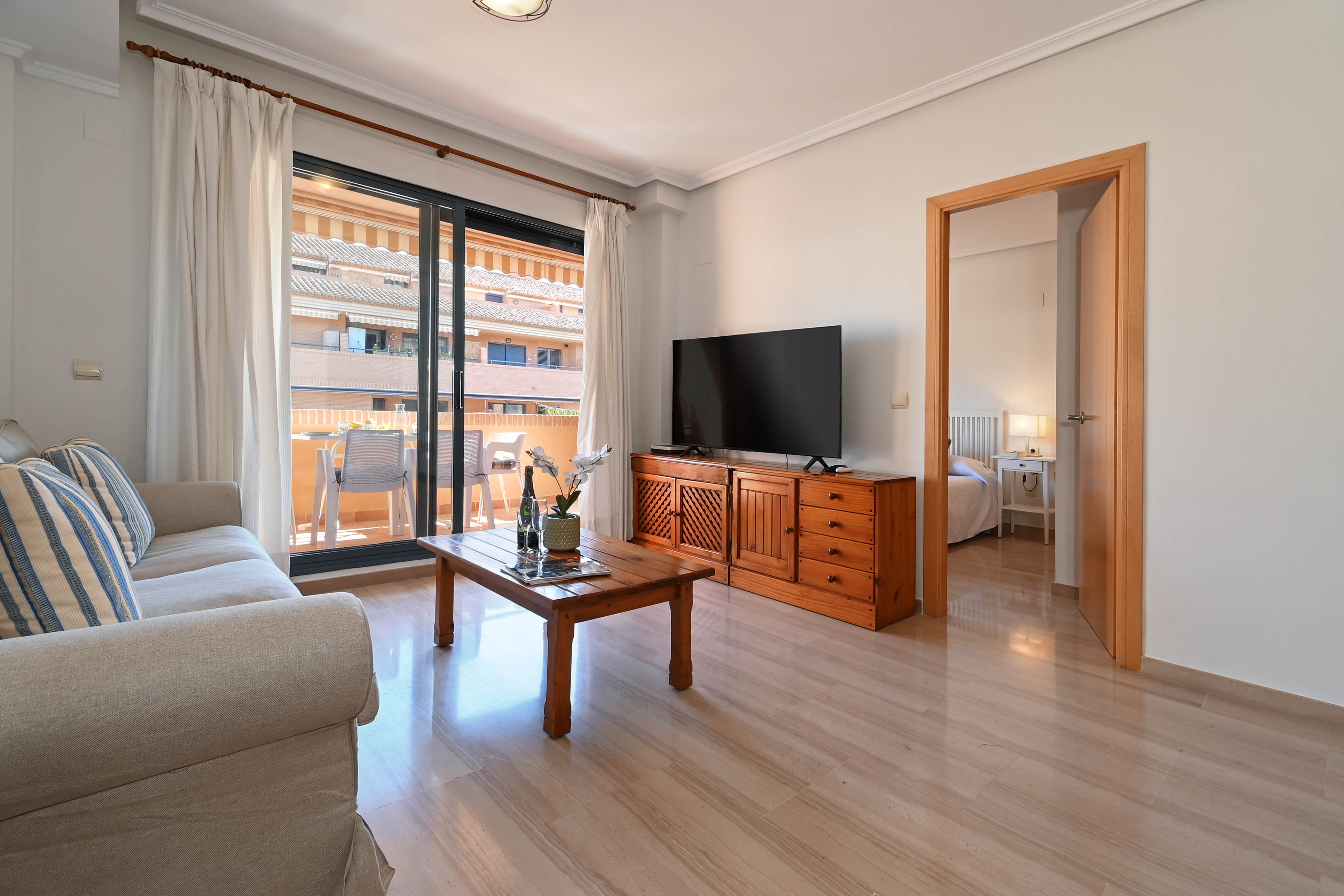 Bright living area featuring polished marble floors, natural wood furniture, and direct access to a furnished terrace through large sliding glass doors.