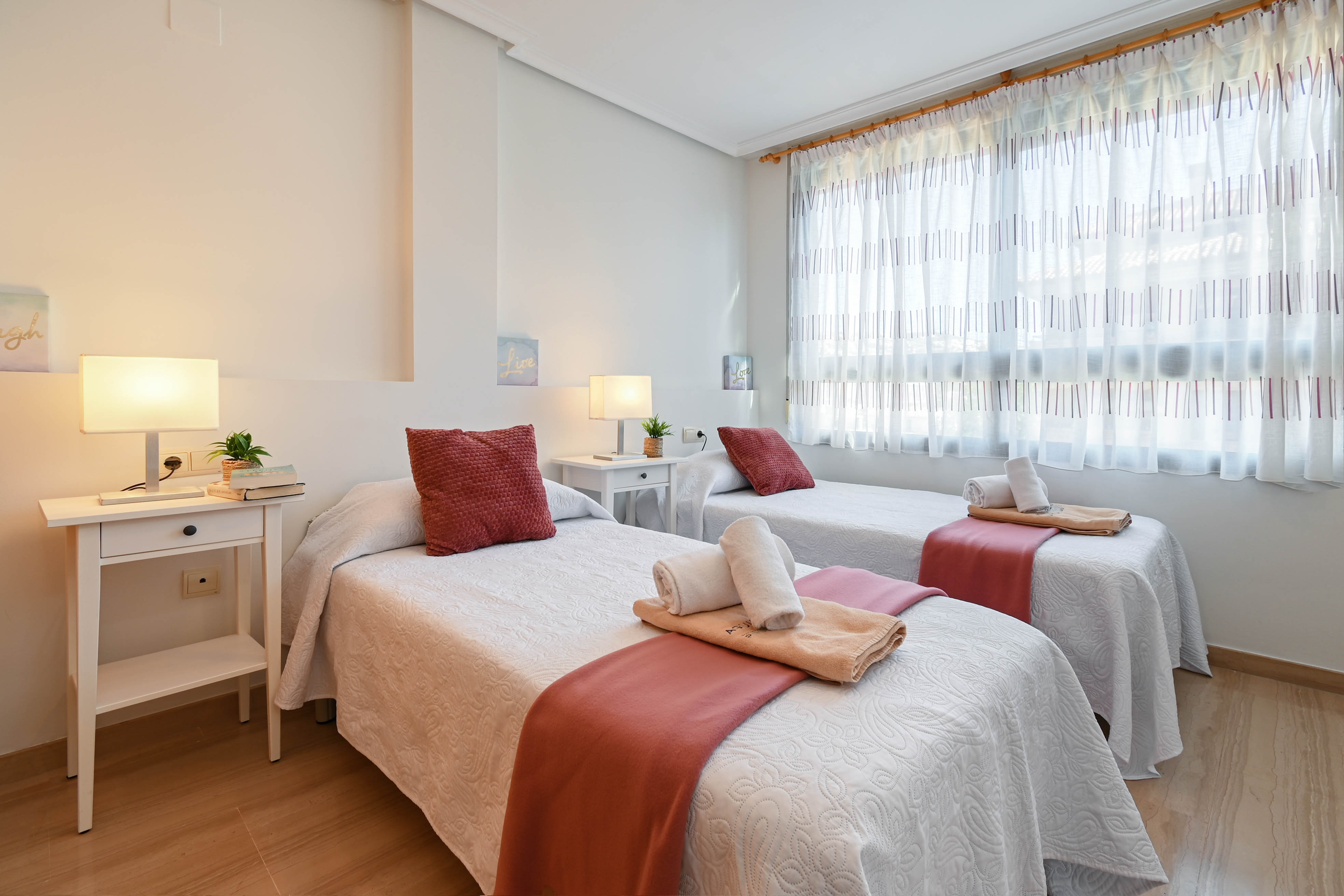 Bright twin bedroom featuring two single beds, white bedside tables, and light wood flooring. A large window provides soft natural light through patterned translucent curtains.