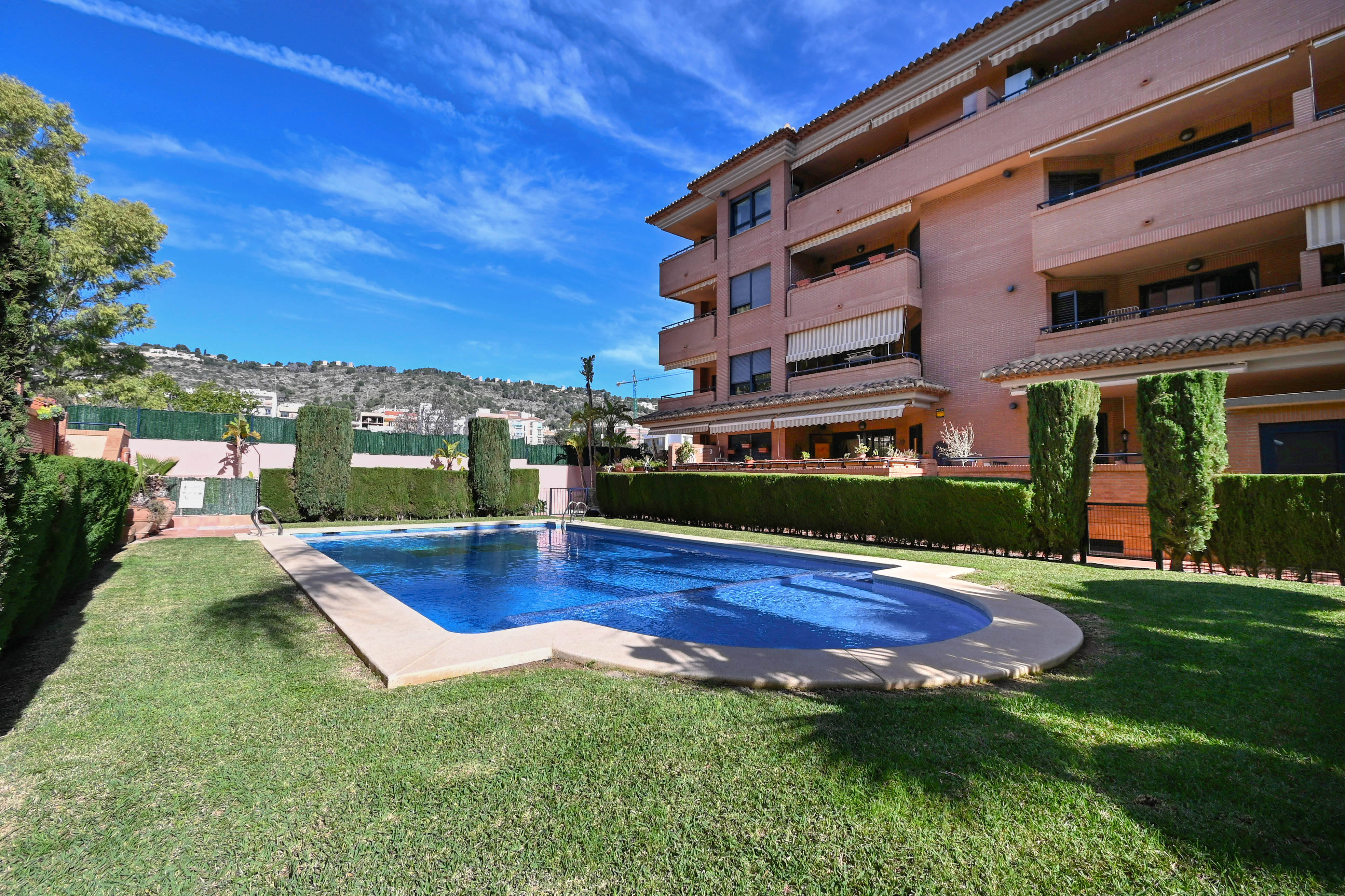 Communal swimming pool with stone coping, set within a manicured lawn and bordered by privacy hedges against a brick apartment complex.