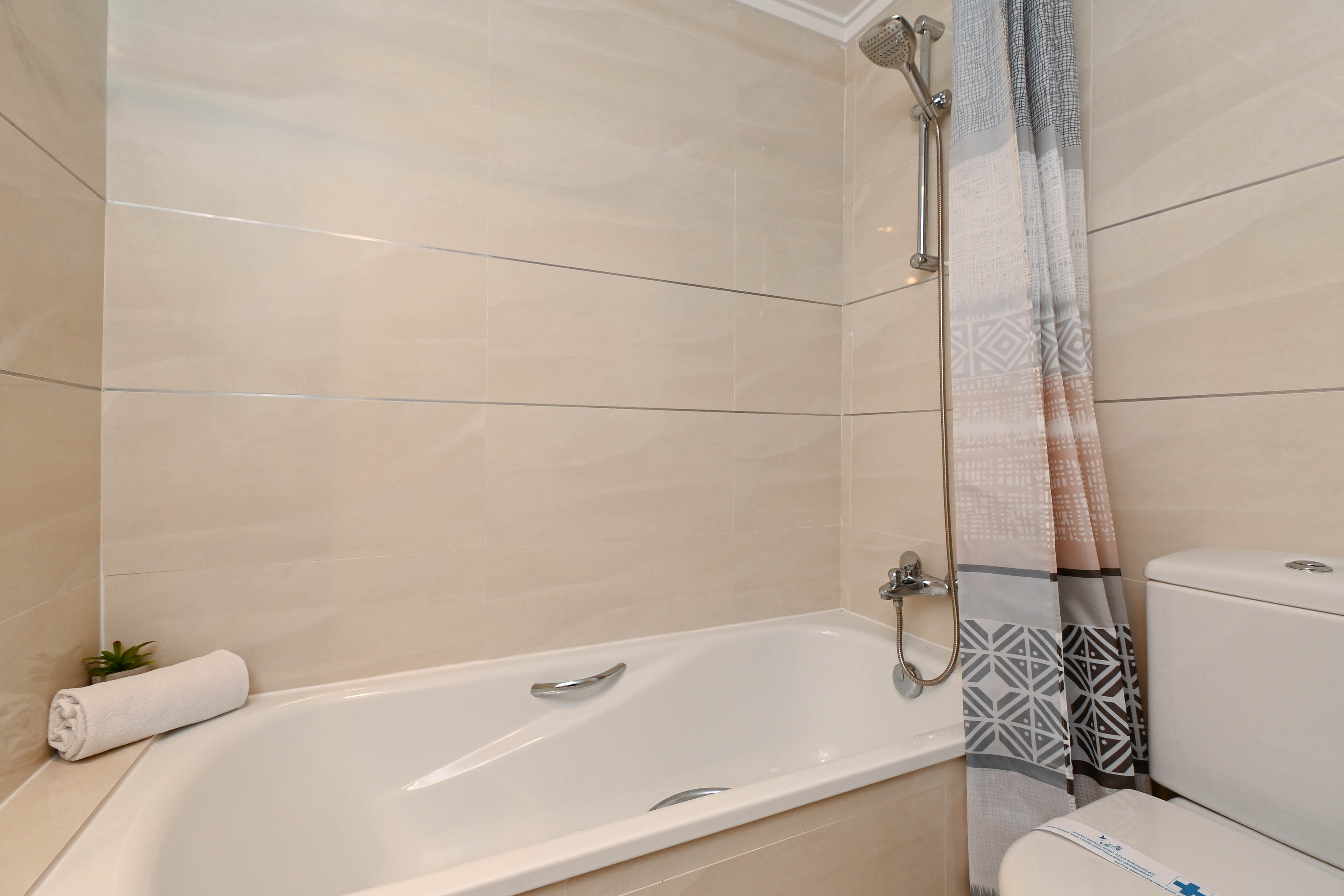 Functional bathroom featuring a bathtub with chrome shower fixtures, beige tiled walls with horizontal accents, and a geometric patterned shower curtain.