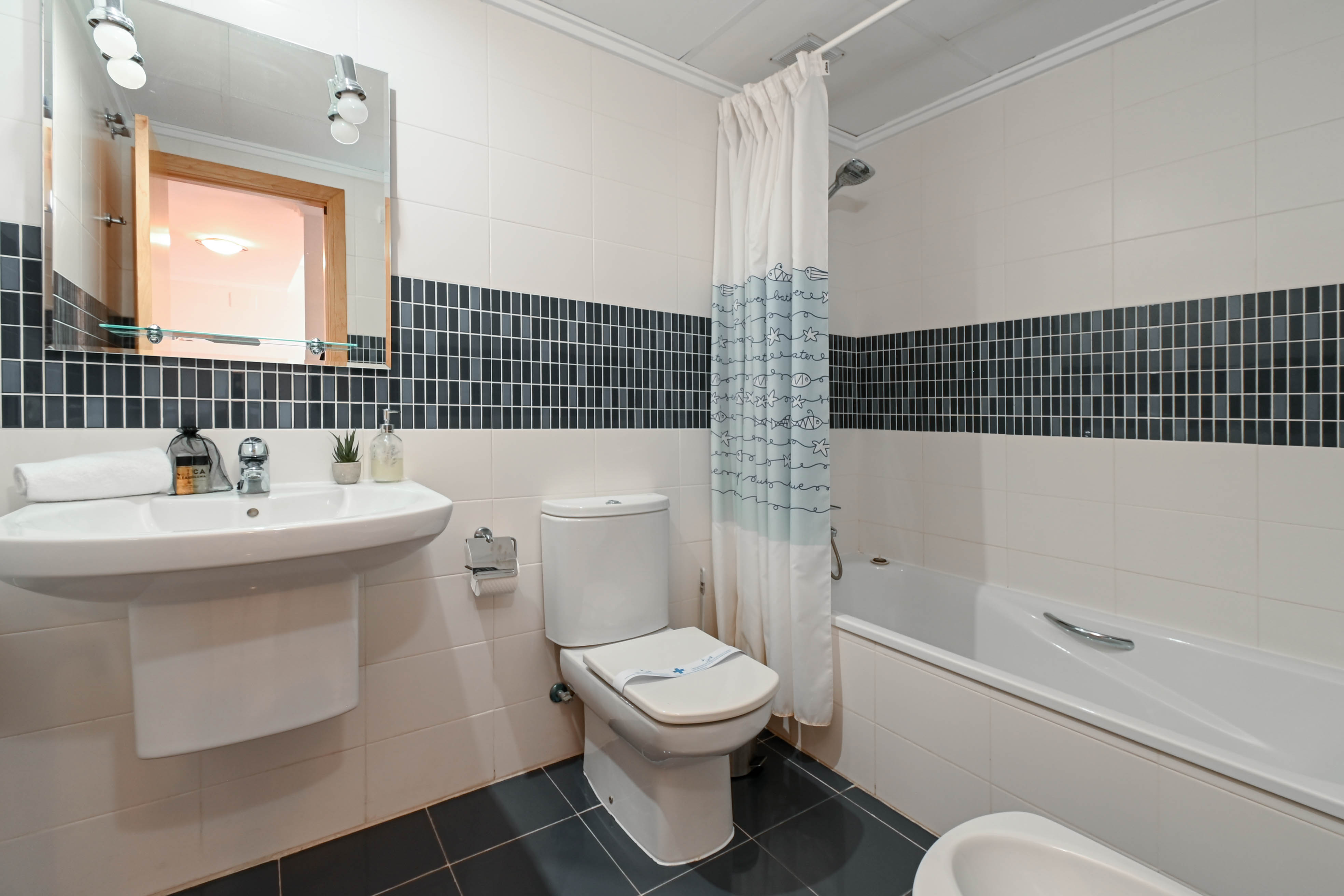 Functional bathroom with white wall tiles and a dark mosaic accent, featuring a bathtub, pedestal sink, and mirror with integrated lighting.