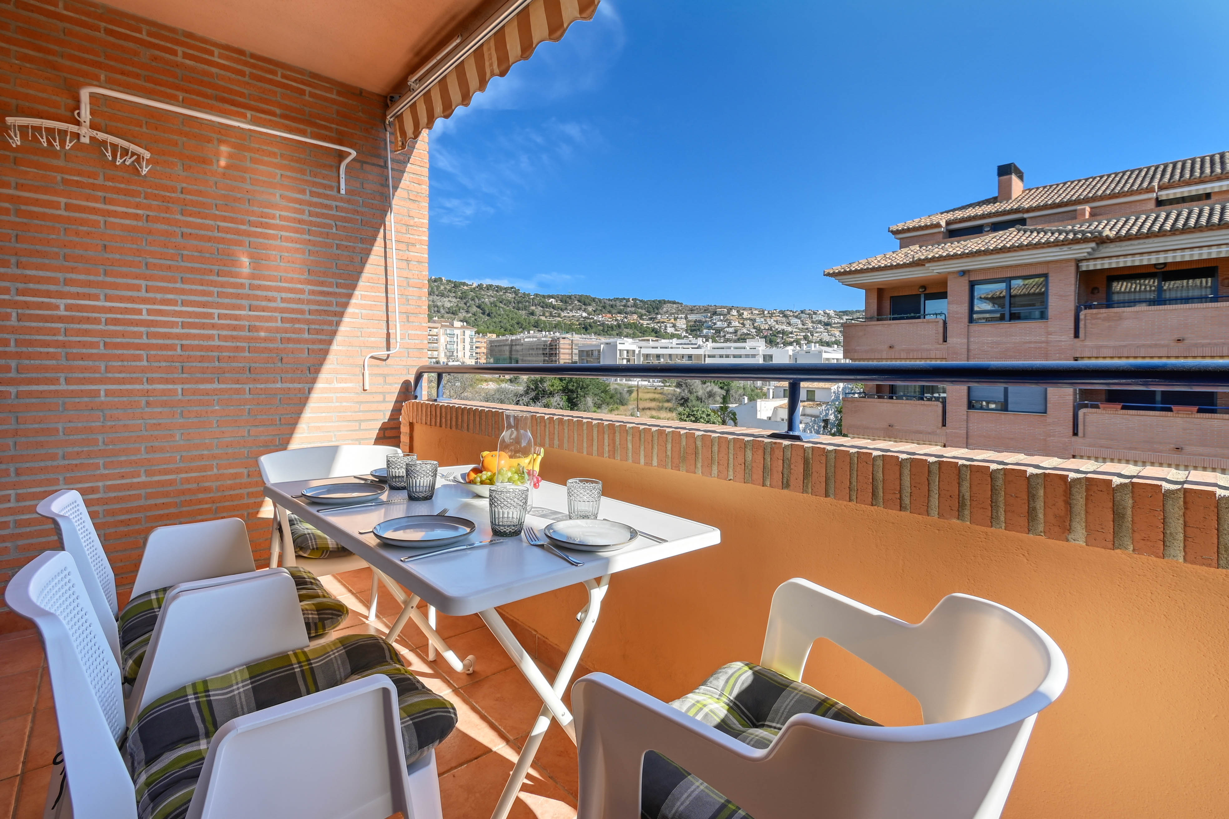 Private terrace with an outdoor dining table, striped awning, and brick walls. It features clear views of the surrounding hills and the residential area of Jávea.