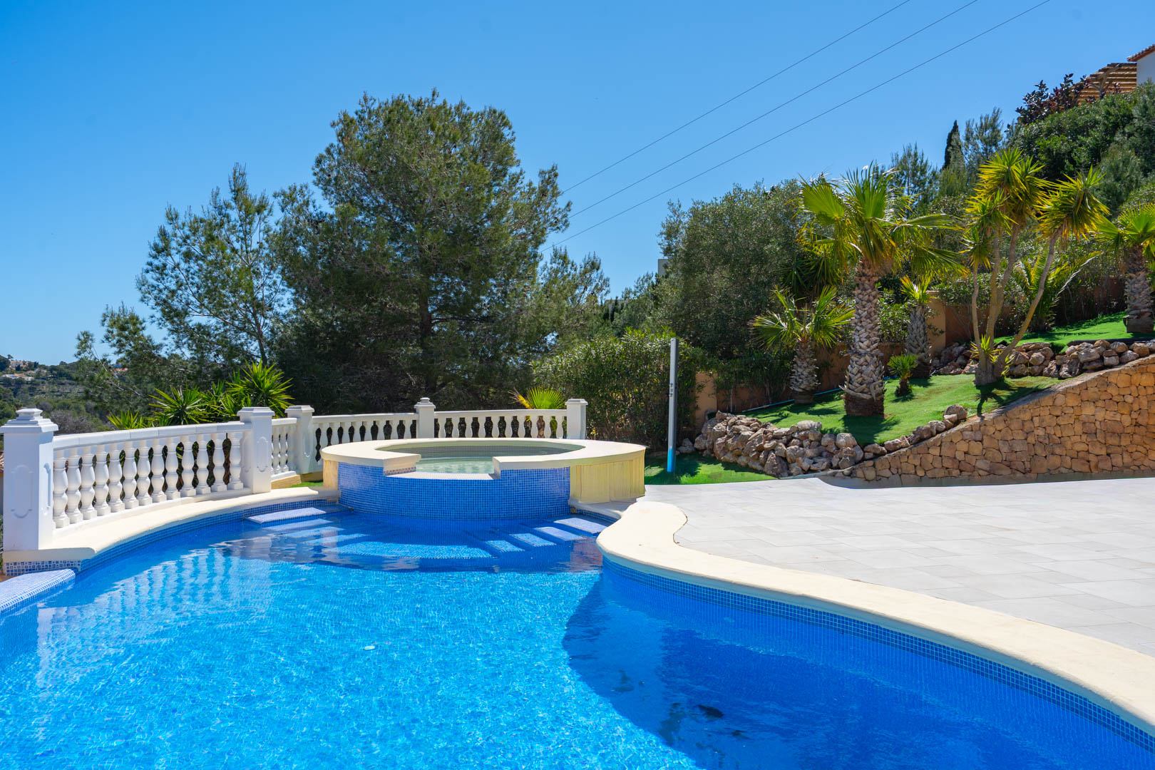A 10x5 m swimming pool featuring an integrated circular spa, blue mosaic tiling, a white stone balustrade, and a landscaped garden with palm trees.