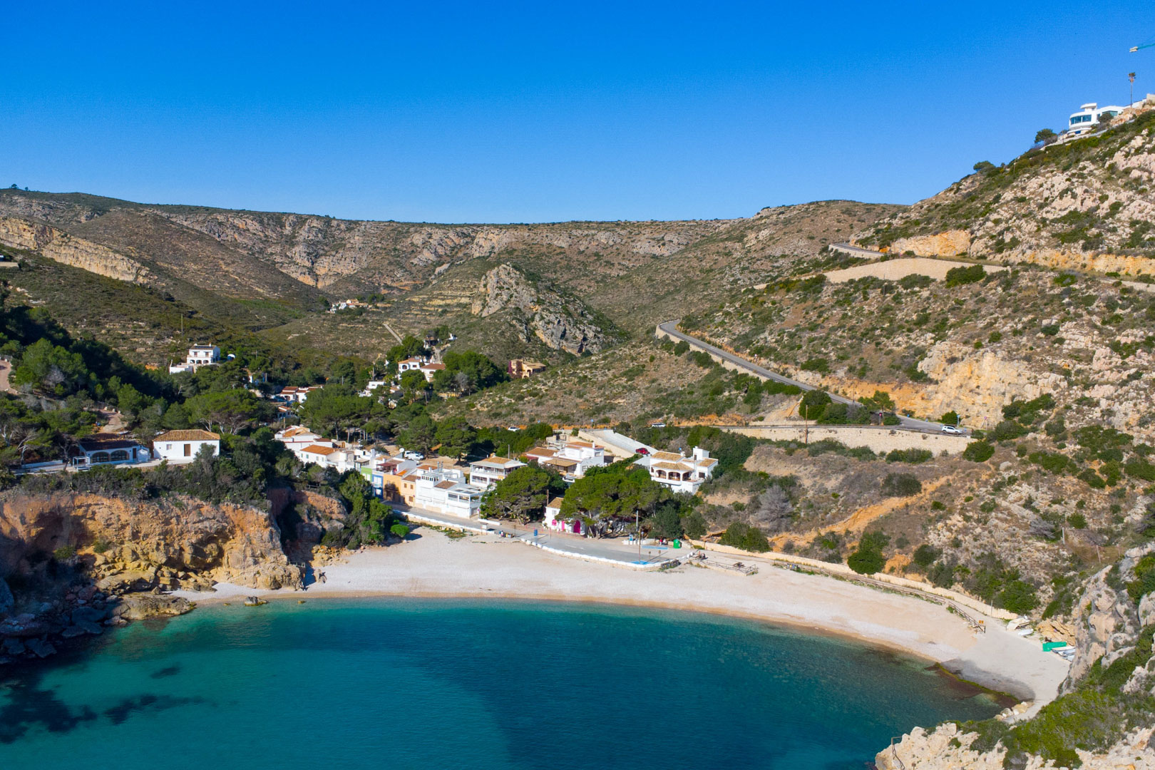 Aerial view of Cala Granadella in Jávea, featuring turquoise waters, a pebble beach, and white houses nestled between rugged Mediterranean cliffs.