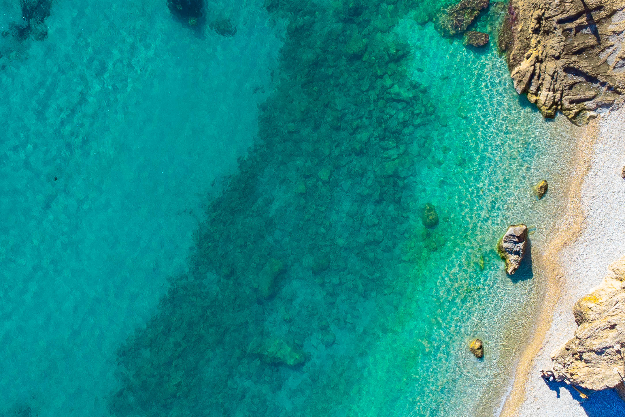 Aerial view of the Jávea coastline featuring crystal-clear turquoise waters, a visible rocky seabed, and a natural pebble shoreline.