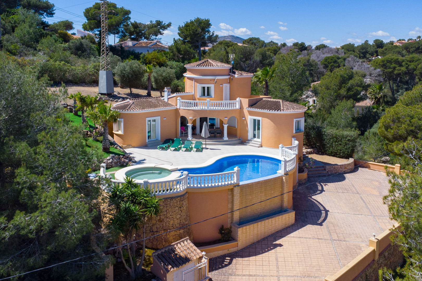 Aerial view of the Mediterranean villa featuring a curved swimming pool with integrated spa, white balustrades, and a large paved driveway surrounded by pine trees.