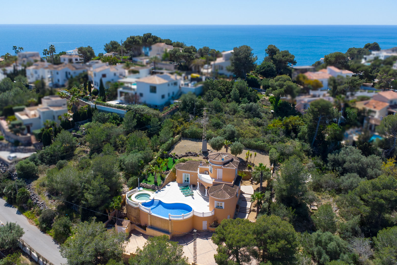 Aerial view of the villa featuring a terracotta facade, kidney-shaped pool with white balustrades, and an elevated position offering sea views amidst Mediterranean greenery.