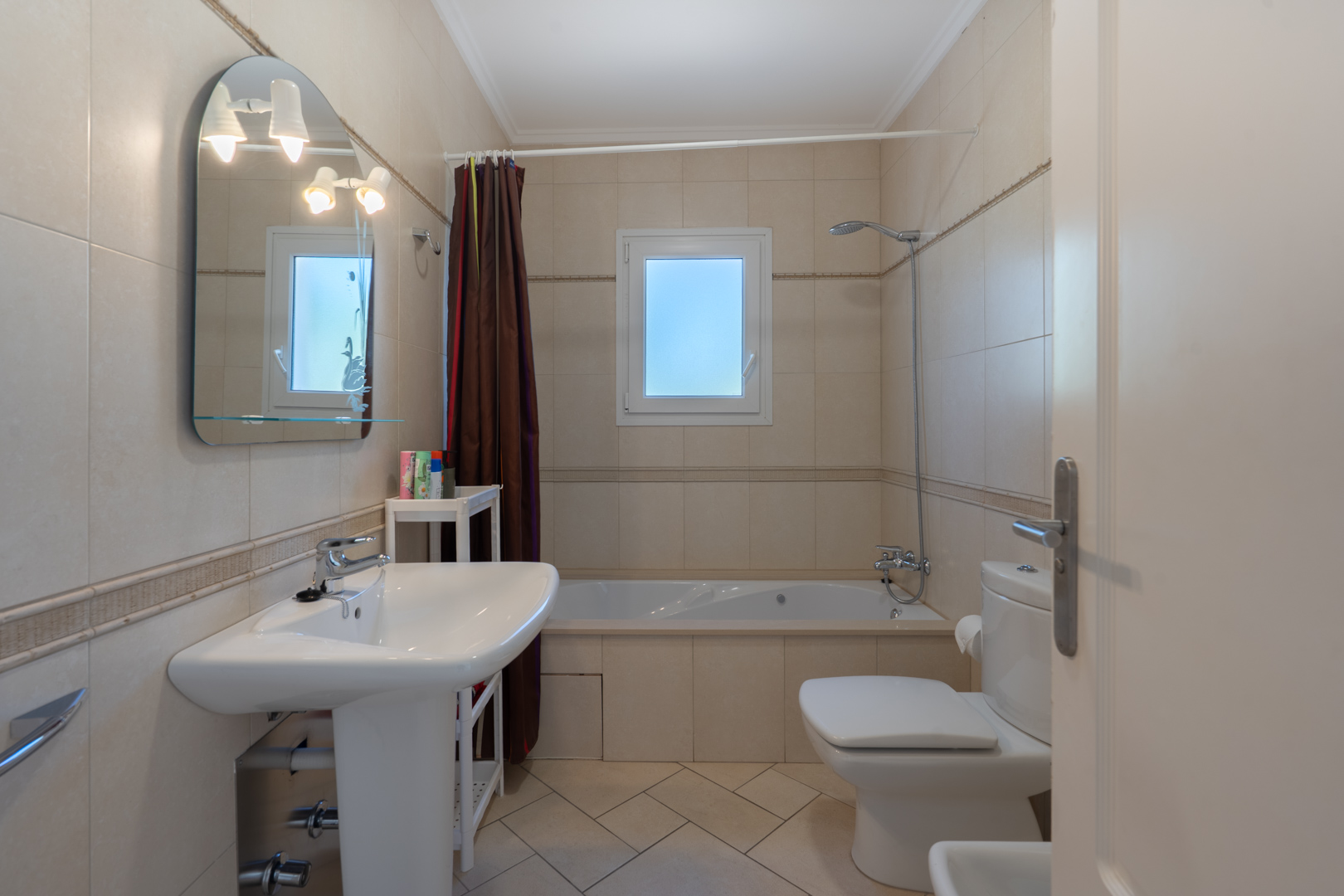 Bathroom featuring beige wall tiles, a built-in bathtub, a pedestal sink, and an arched mirror with integrated lighting next to a frosted PVC window.