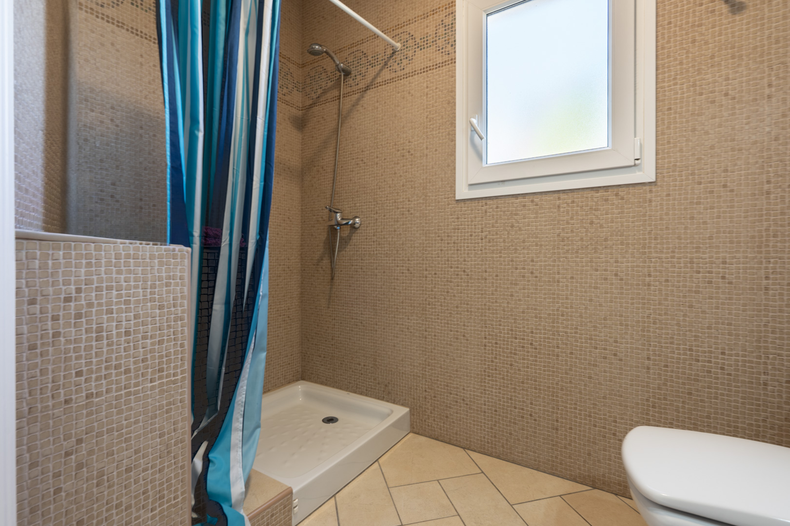 Bathroom featuring brown mosaic wall tiles, a white shower tray with a striped curtain, and a white PVC window providing natural light and ventilation.