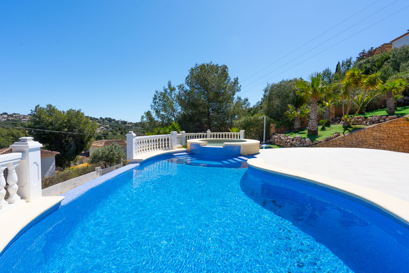 Blue mosaic pool with an integrated circular spa, featuring a white balustrade, expansive paved terrace, and landscaped garden with palm trees.