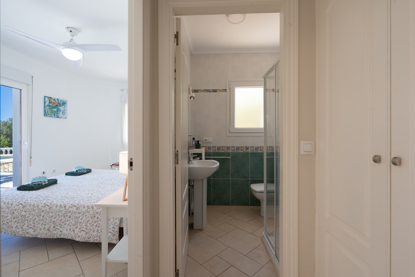 Bright bedroom featuring a ceiling fan and terrace access, adjacent to a bathroom with a glass shower enclosure and hallway built-in wardrobes.