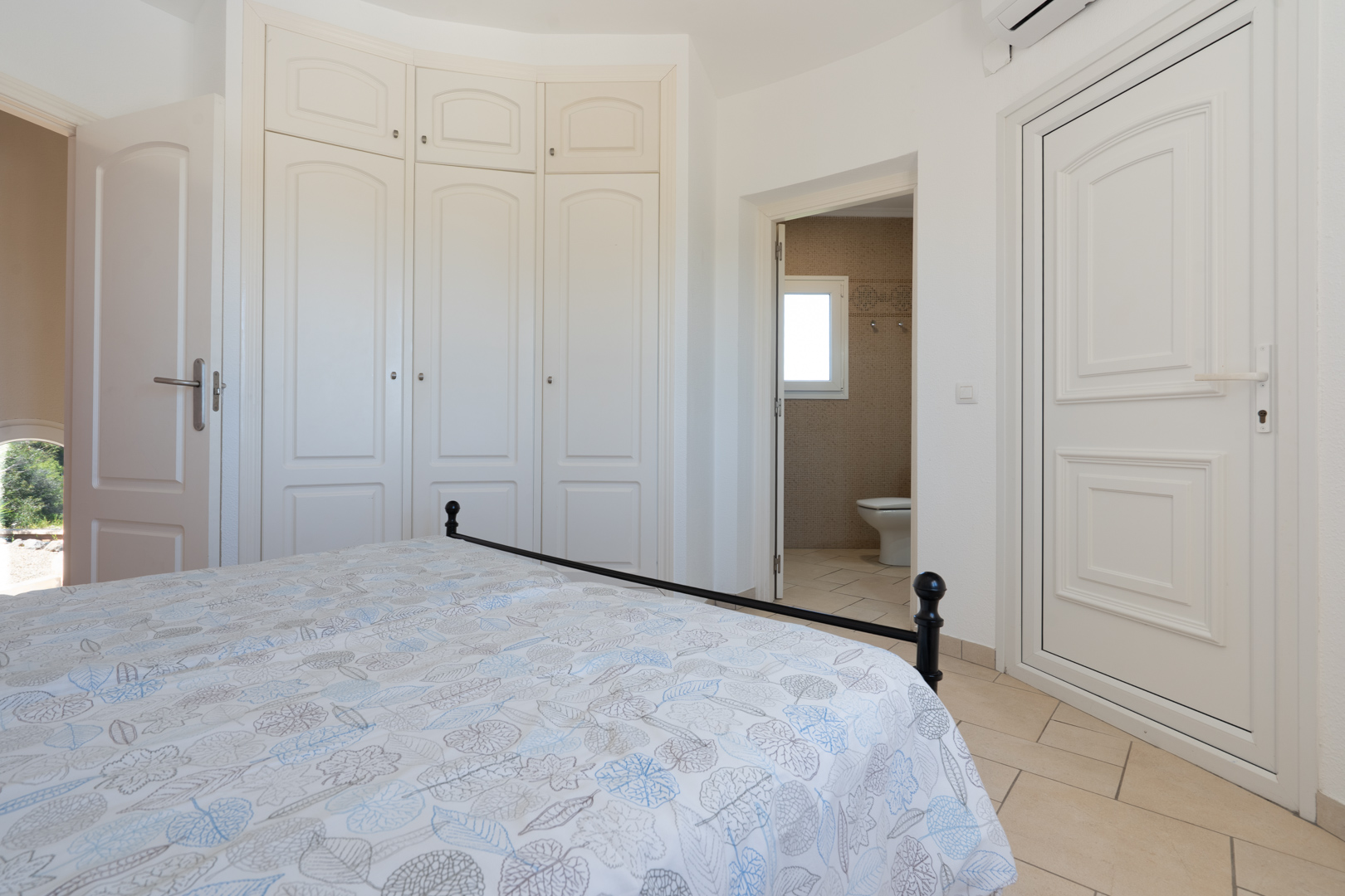 Bright bedroom featuring white built-in wardrobes, ceramic tile flooring, and direct access to an en-suite bathroom with mosaic tiling.