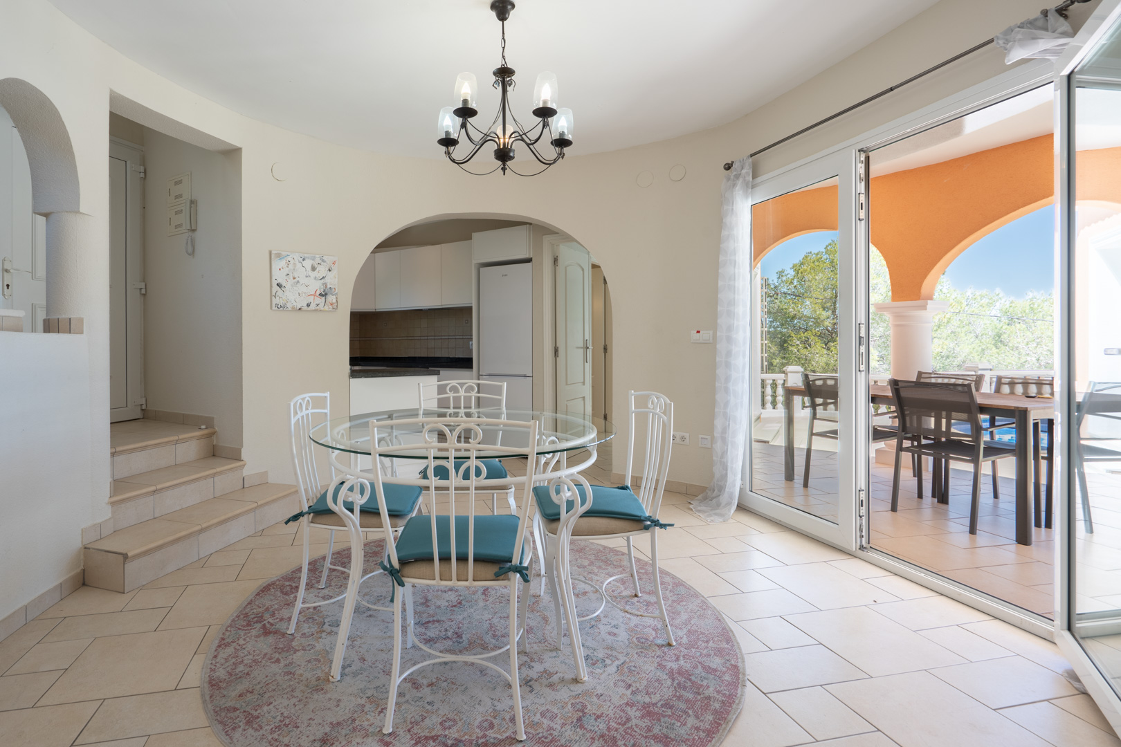Bright dining area featuring a glass-top table and white wrought-iron chairs, with arched access to the kitchen and sliding doors to the terrace.
