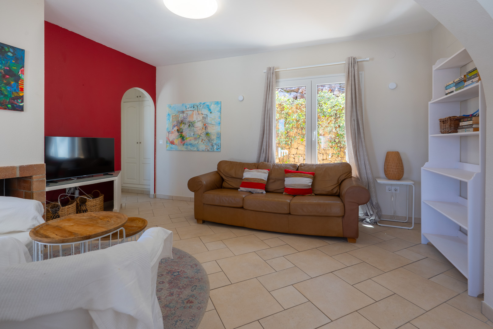 Bright living area featuring a brown leather sofa, red accent wall, and ceramic tile flooring. Large PVC windows provide natural light and views of the garden.