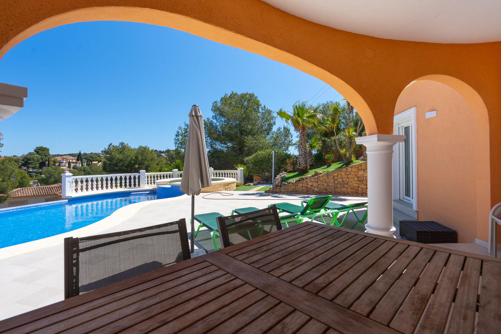 Covered terrace featuring traditional arches and a wooden dining area, overlooking the private pool, sun loungers, and landscaped Mediterranean garden.
