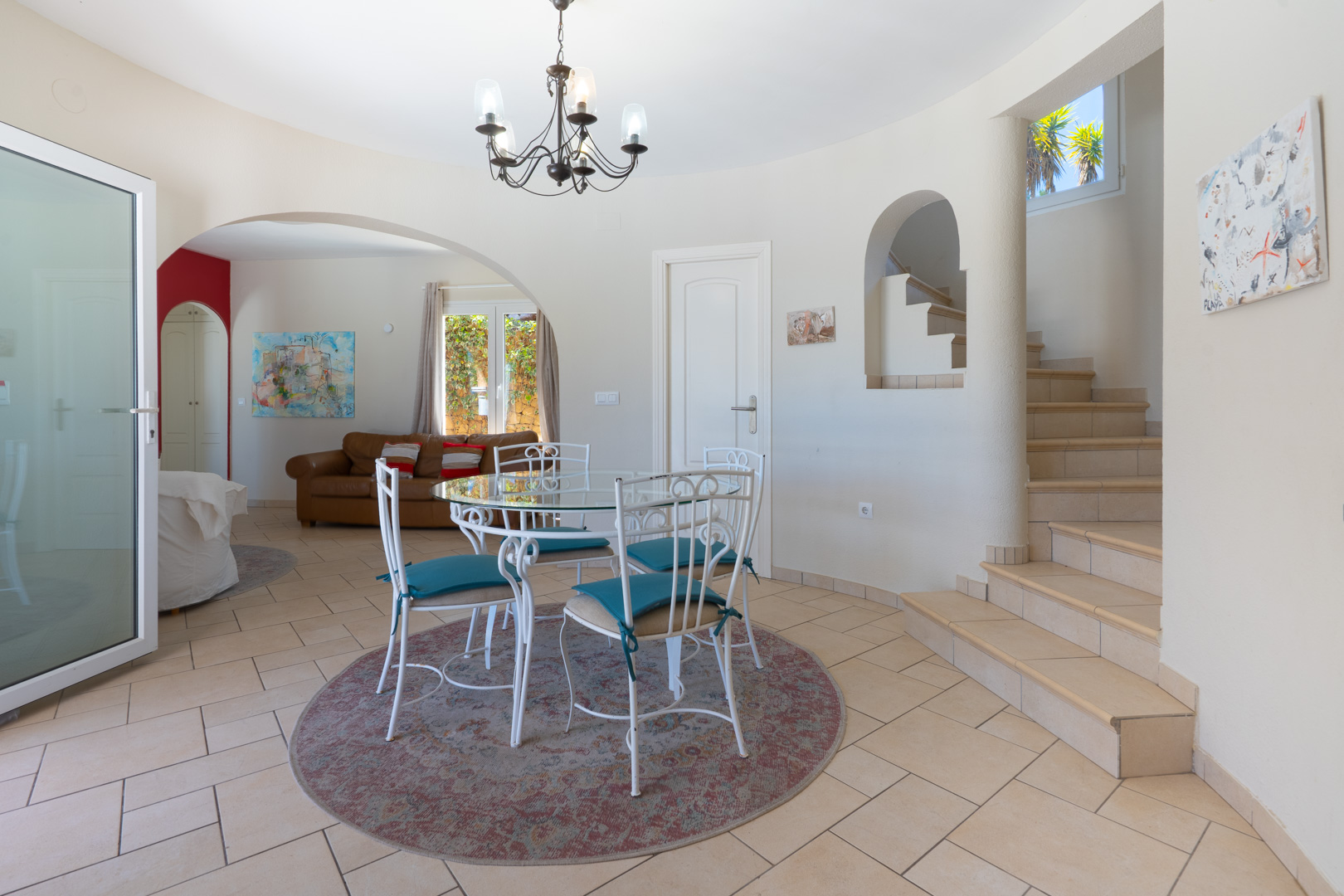 Dining area featuring a glass-top table with white wrought-iron chairs and a chandelier. The space connects to a tiled staircase and an arched living room entrance.
